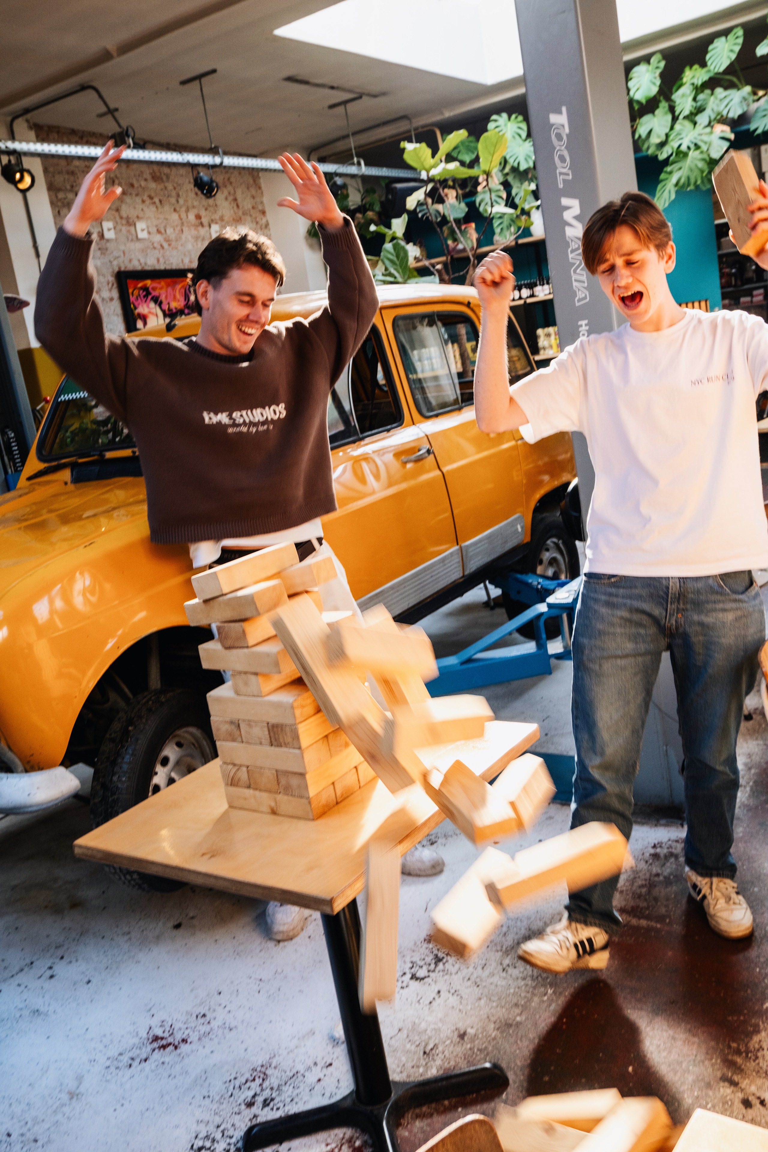 Twee lachende mannen vieren een vallende Jenga-toren in een lichte ruimte met een gele auto op de achtergrond.