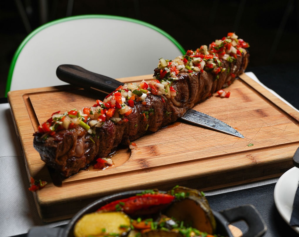 Grilled steak skewer topped with vibrant salsa on a wooden board, knife embedded, cozy dining setting.