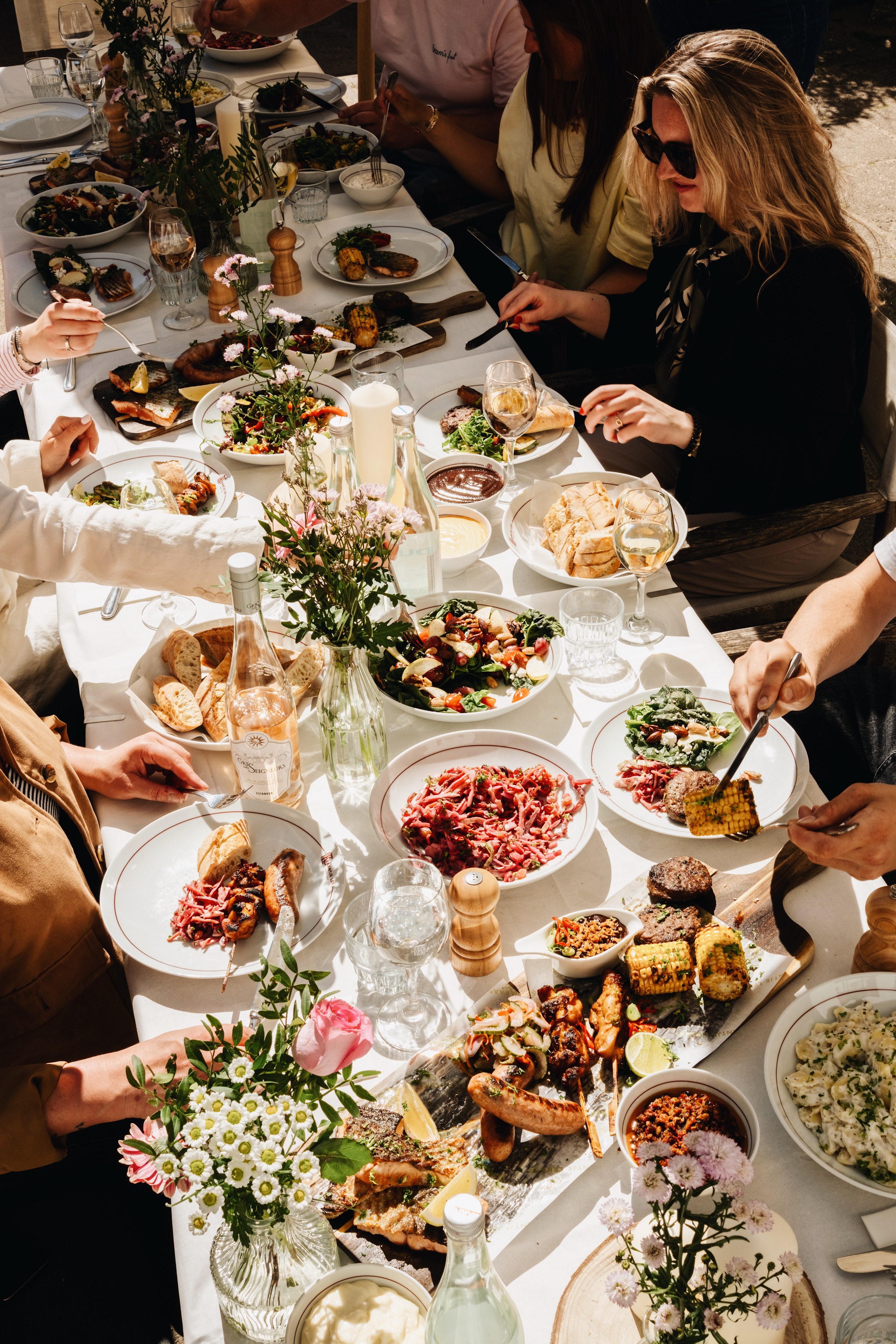 Gezellige lunch met diverse gerechten op een lange tafel, omringd door mensen, bloemen en zonlicht.