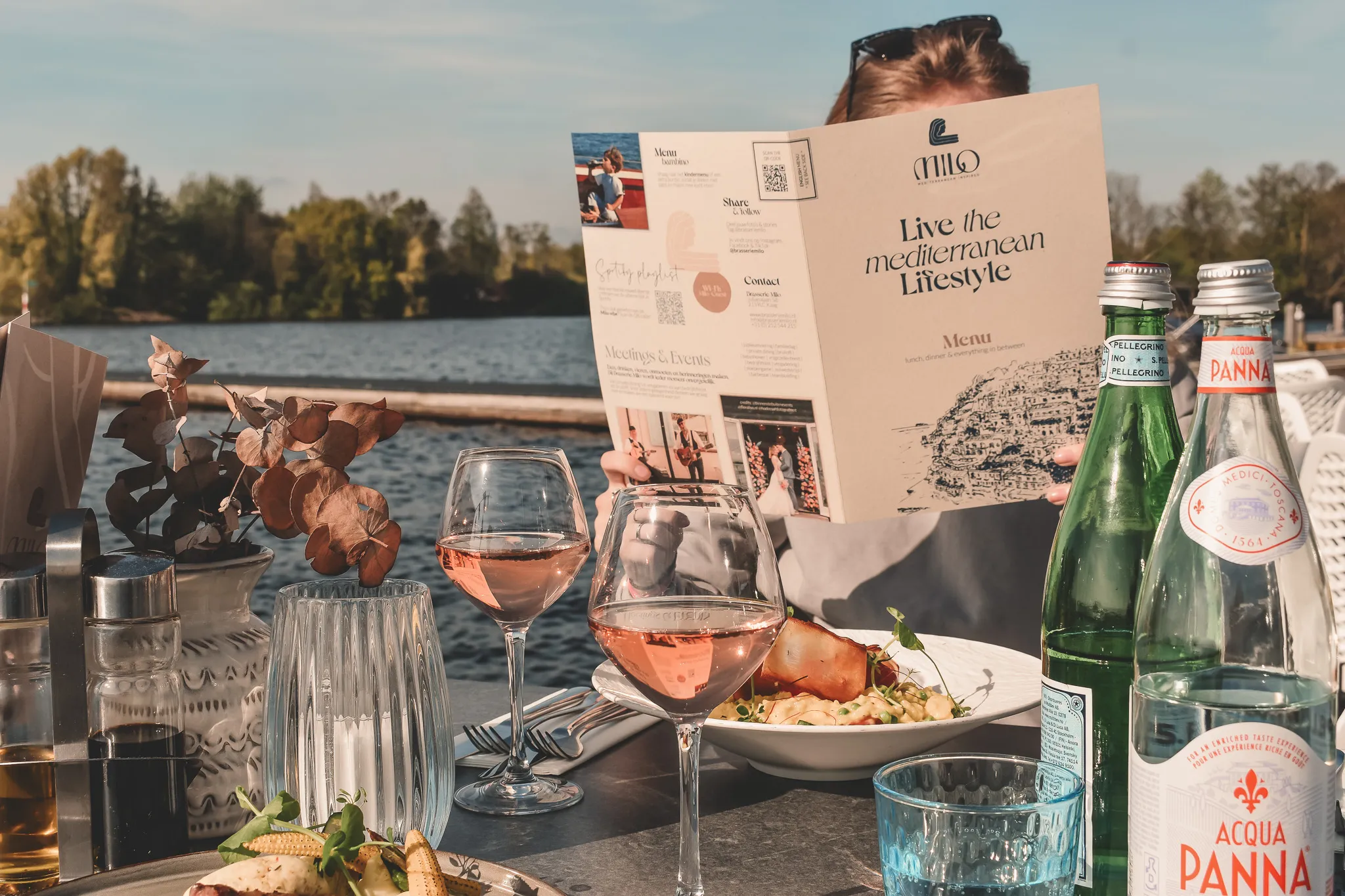 Tafel aan het water met glazen rosé, flessen water, en een menu, omgeven door groene bomen en blauwe lucht.