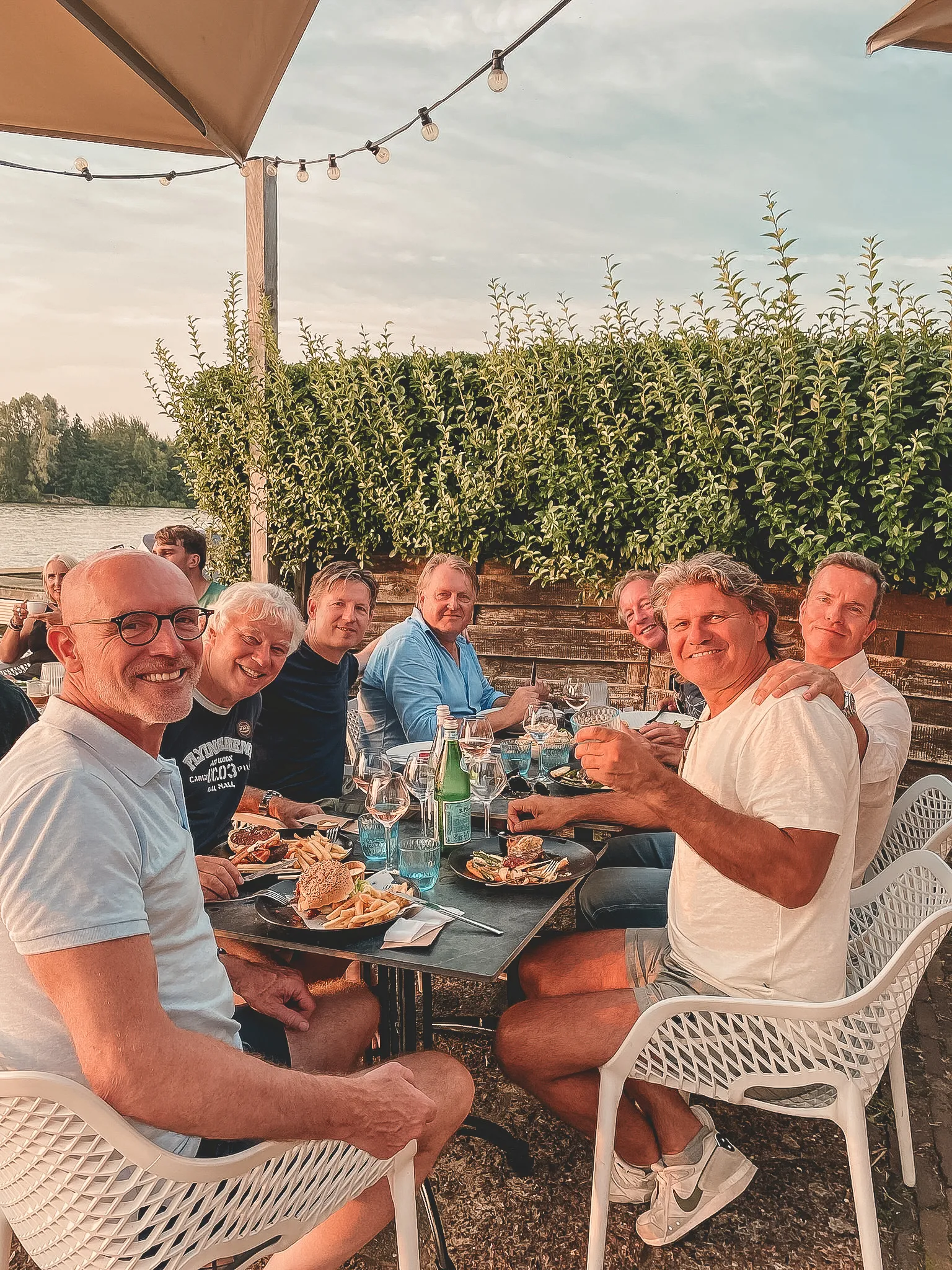 Groep mannen geniet van hamburgers en frietjes op een terras met uitzicht op een groene haag en water.