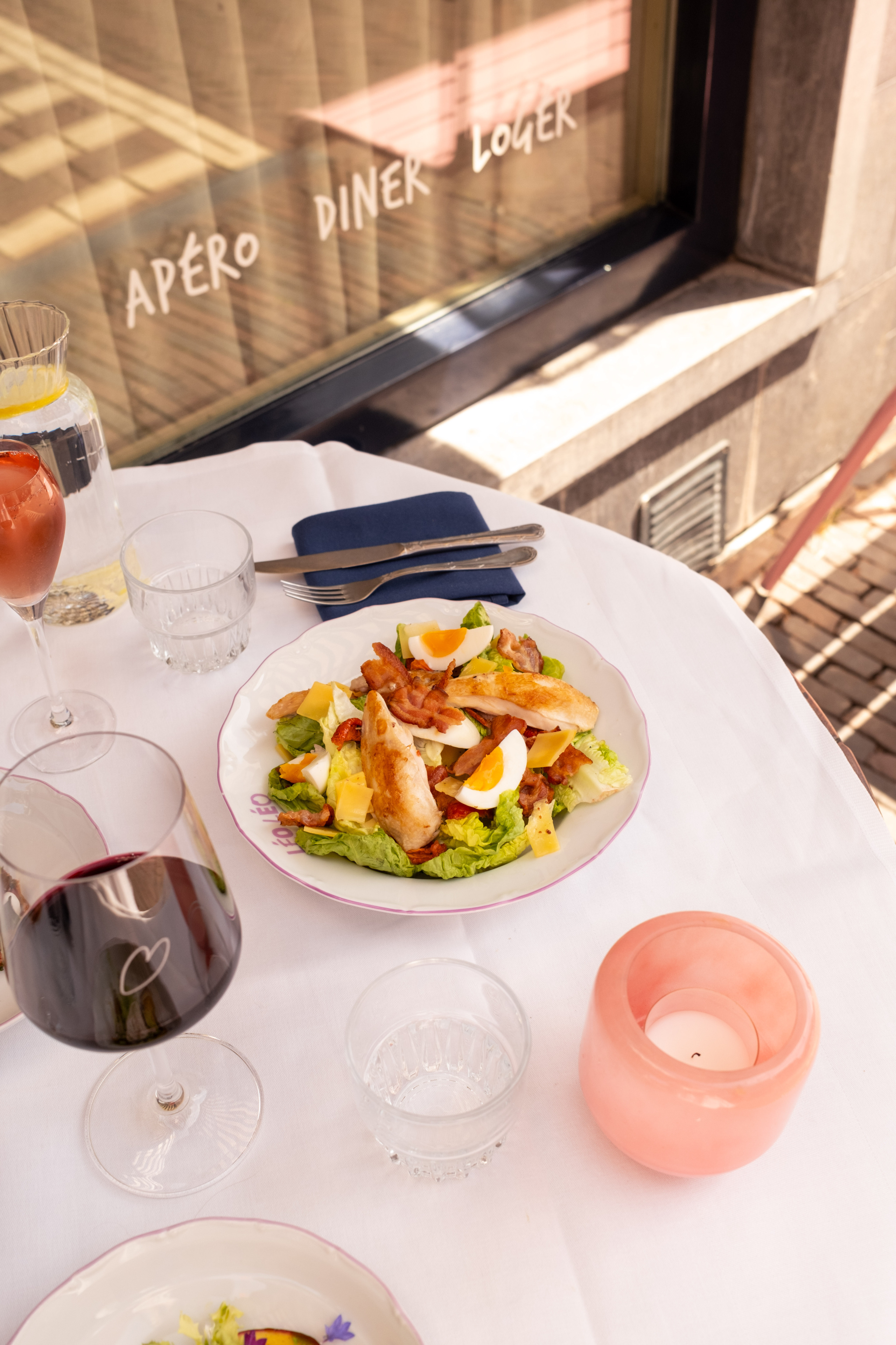 Gekleurde salade met kip, spek en ei op wit tafelkleed, omringd door wijnglazen en een roze kaars.