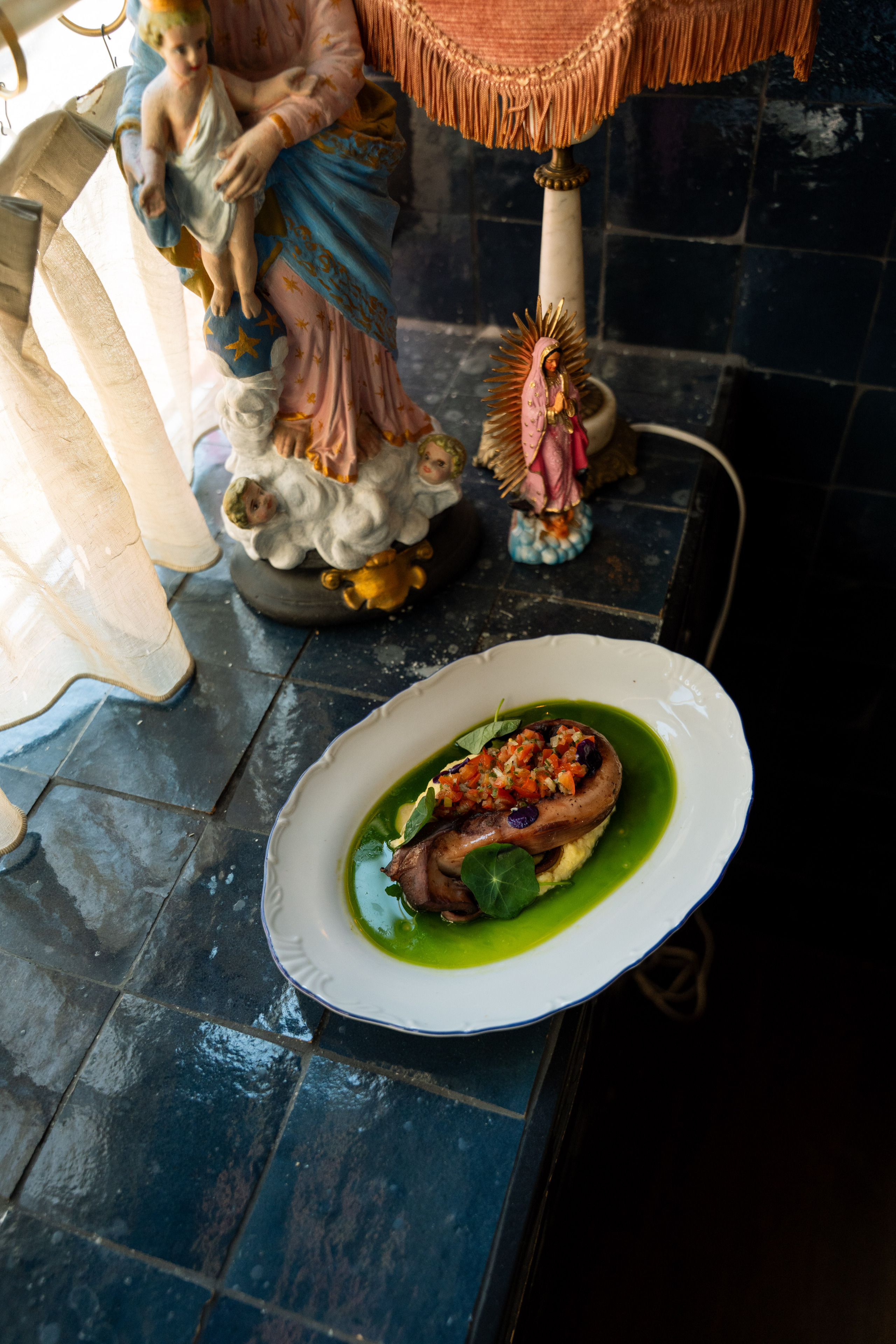 Geroosterde octopus met kleurrijke salsa op een witte schaal, naast religieuze beeldjes op een donkere tegelvloer.