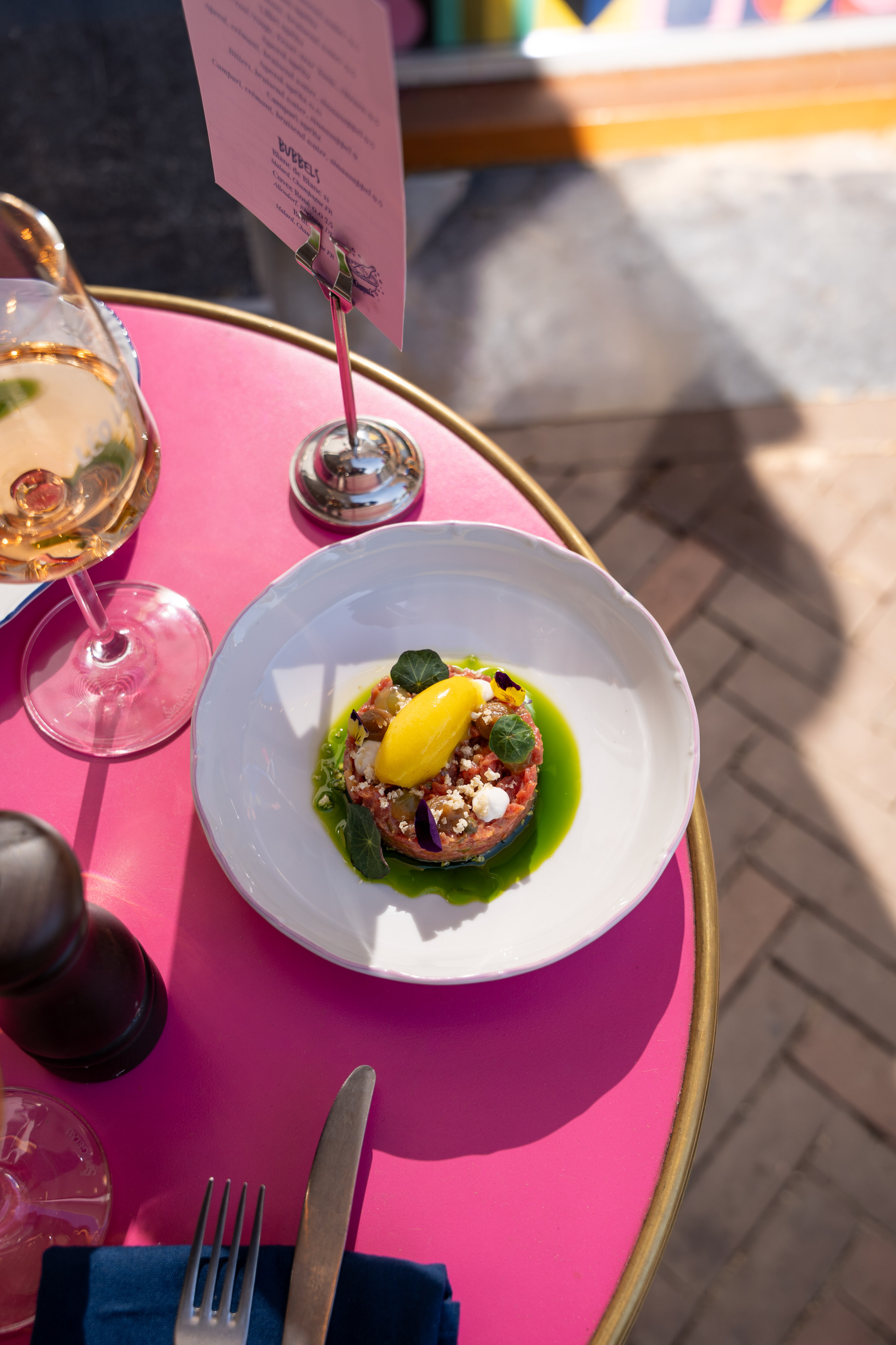 Rundtartaar met gele sorbet en kruiden op een wit bord, op een roze tafel met wijnglas en menukaart.