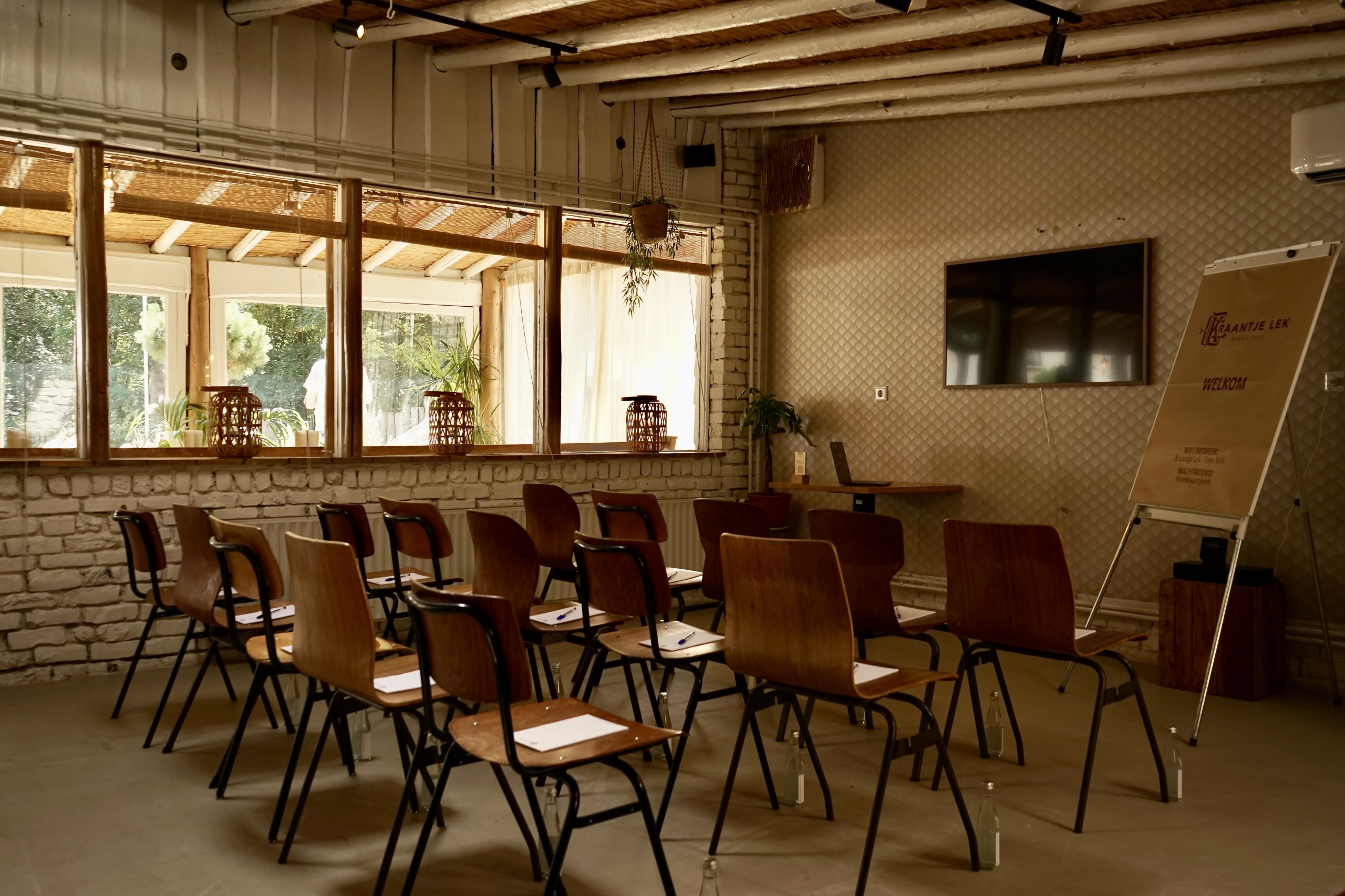 Houten stoelen in een lichte vergaderruimte met grote ramen, planten en een flip-over. Warme, uitnodigende sfeer.