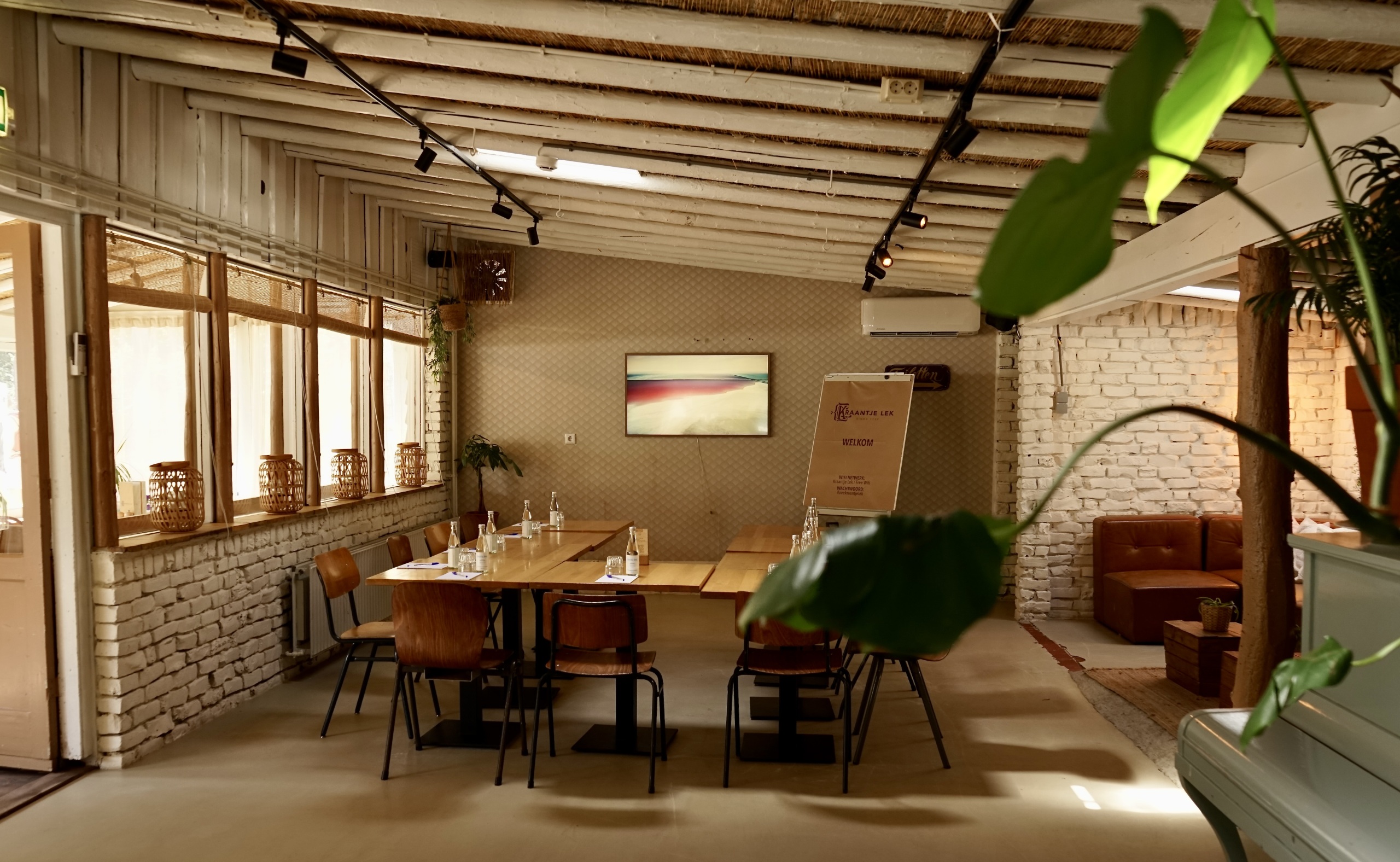 Houten tafel met stoelen in een lichte, rustieke ruimte met witte bakstenen muren en groene planten.
