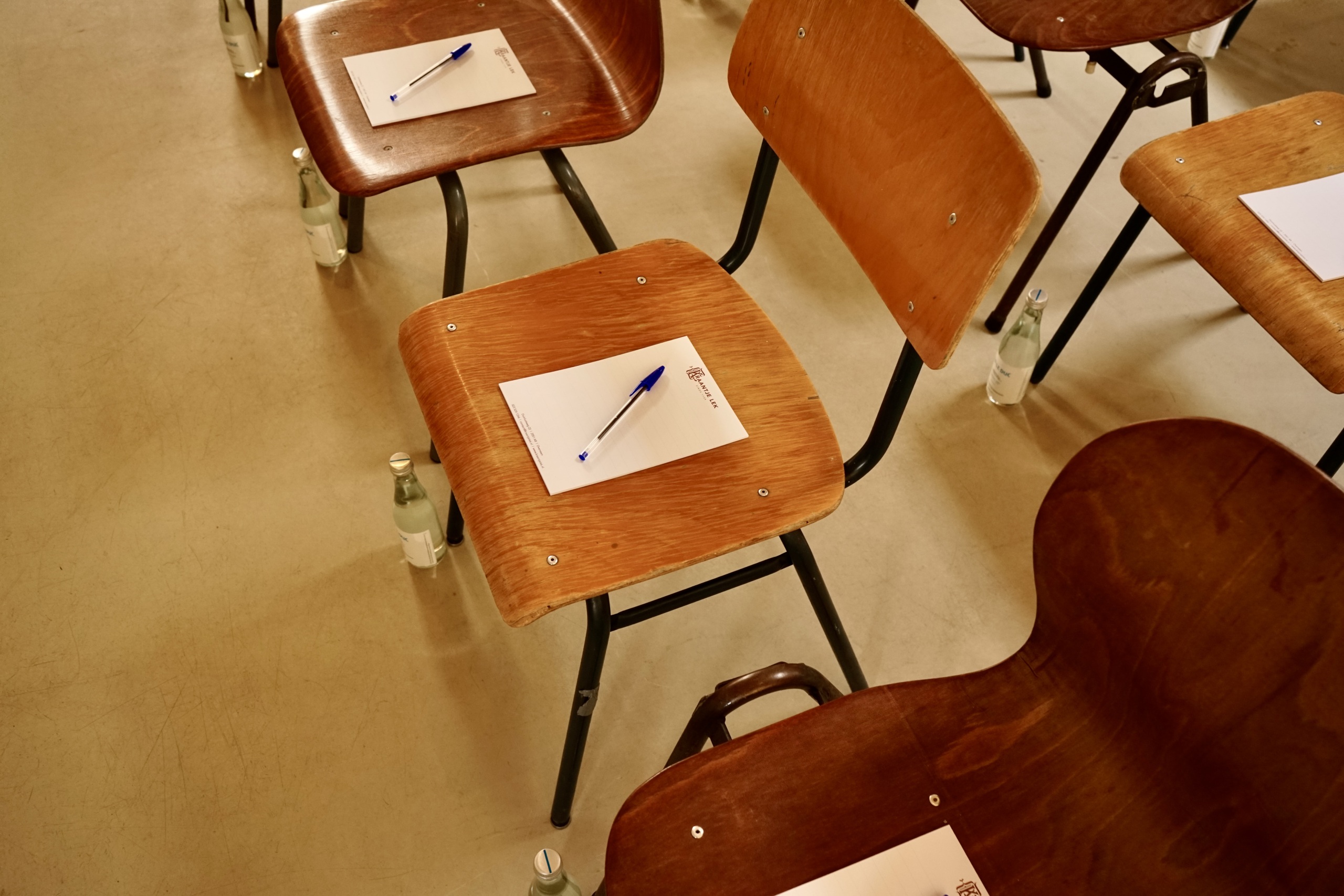 Houten stoelen met schrijfblokken en pennen, naast elke stoel een flesje water op een lichte vloer.
