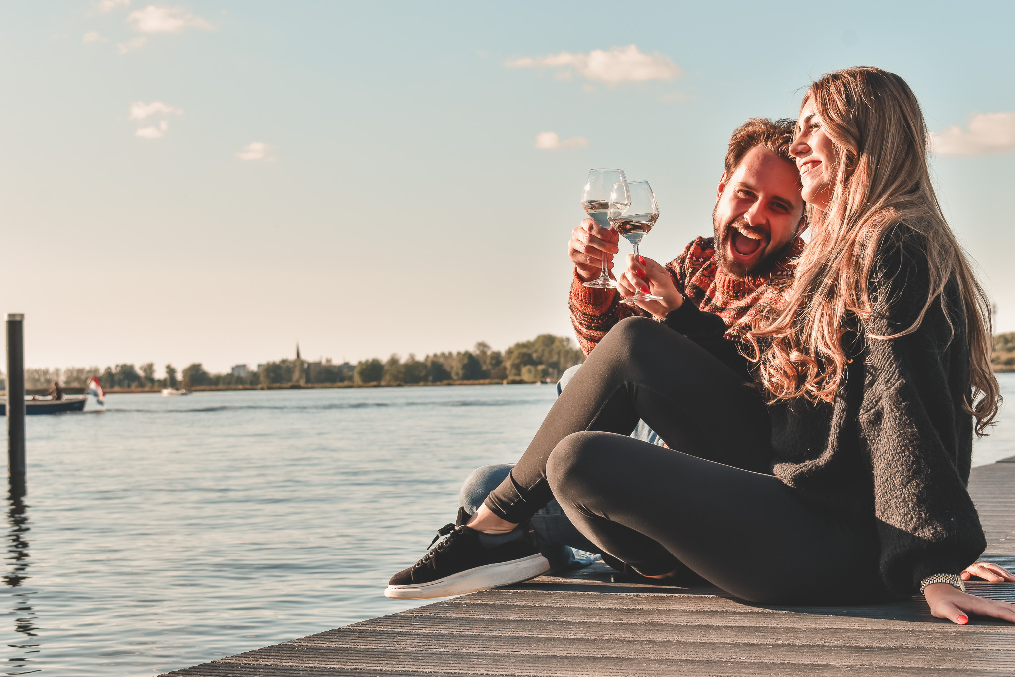 Twee mensen genieten van wijn op een zonnige steiger aan het water, met een blauwe lucht en bomen op de achtergrond.