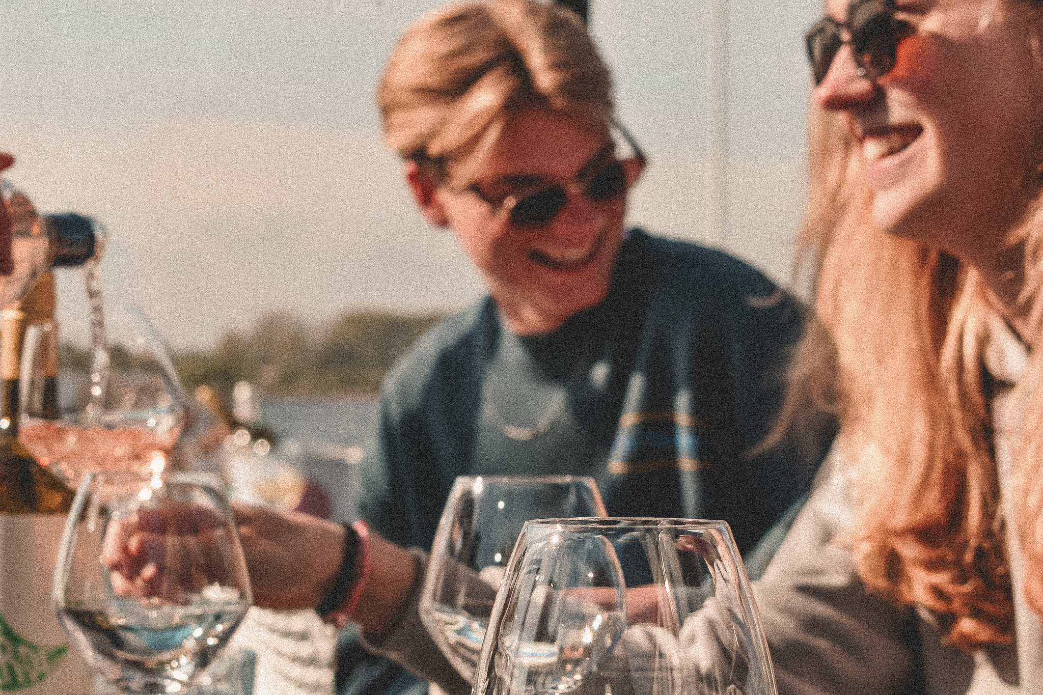 Twee mensen lachen buiten terwijl rosé wijn in glazen wordt geschonken, zonnige sfeer met water op de achtergrond.