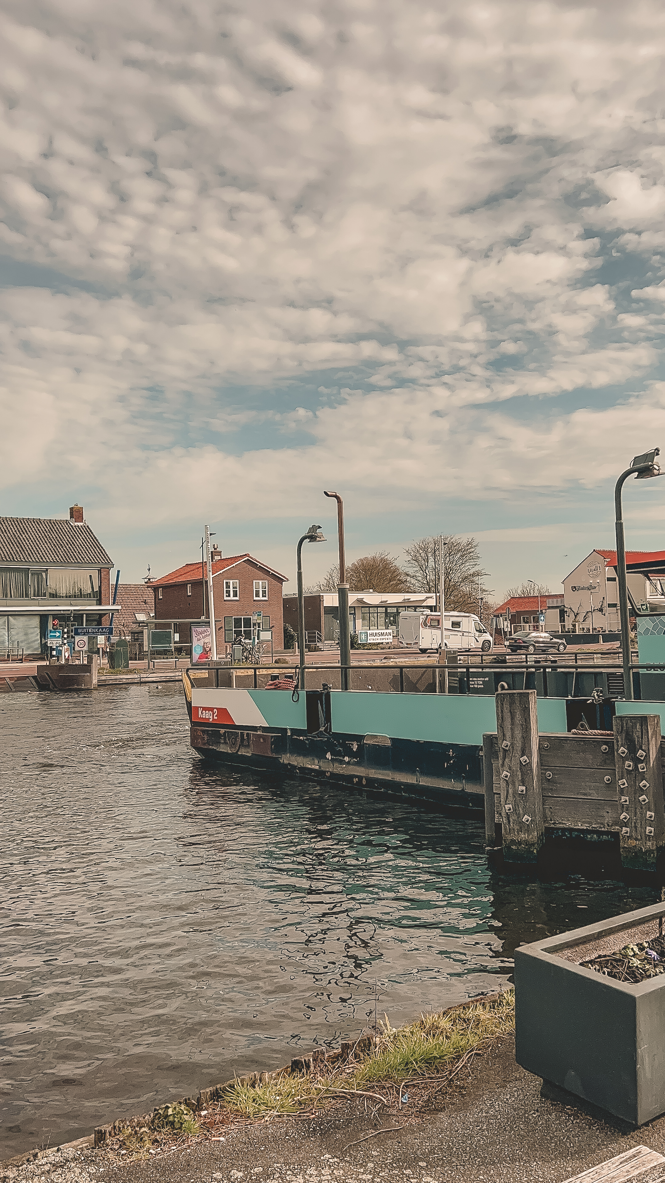 Veerpont op rustig water met blauwe lucht en wolken. Omringd door bakstenen huizen en een camper.