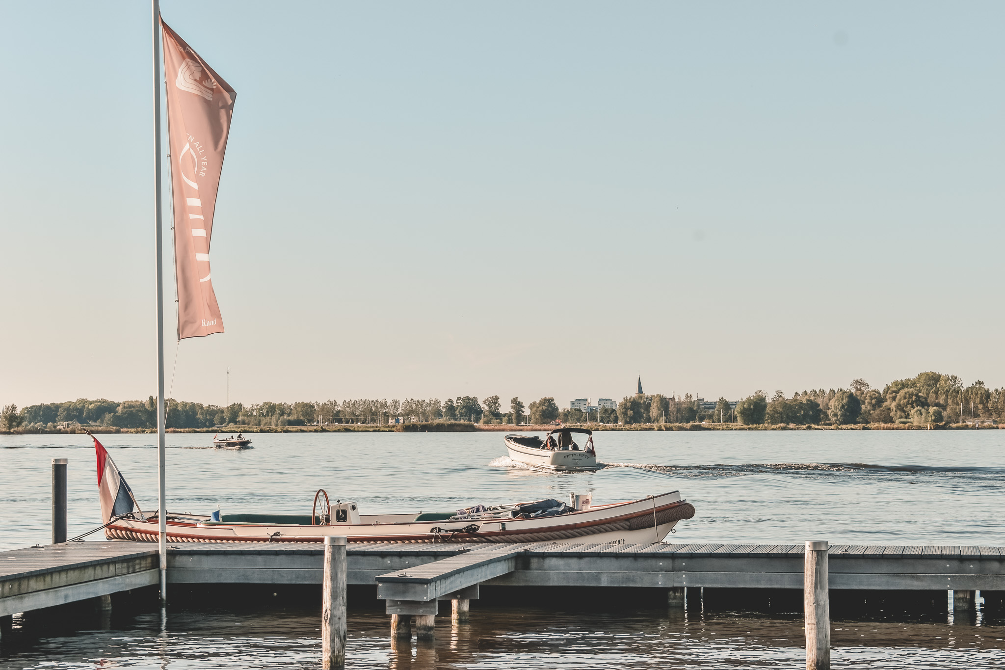 Een rustige steiger met een boot aangemeerd, een andere boot vaart op het meer onder een heldere hemel.