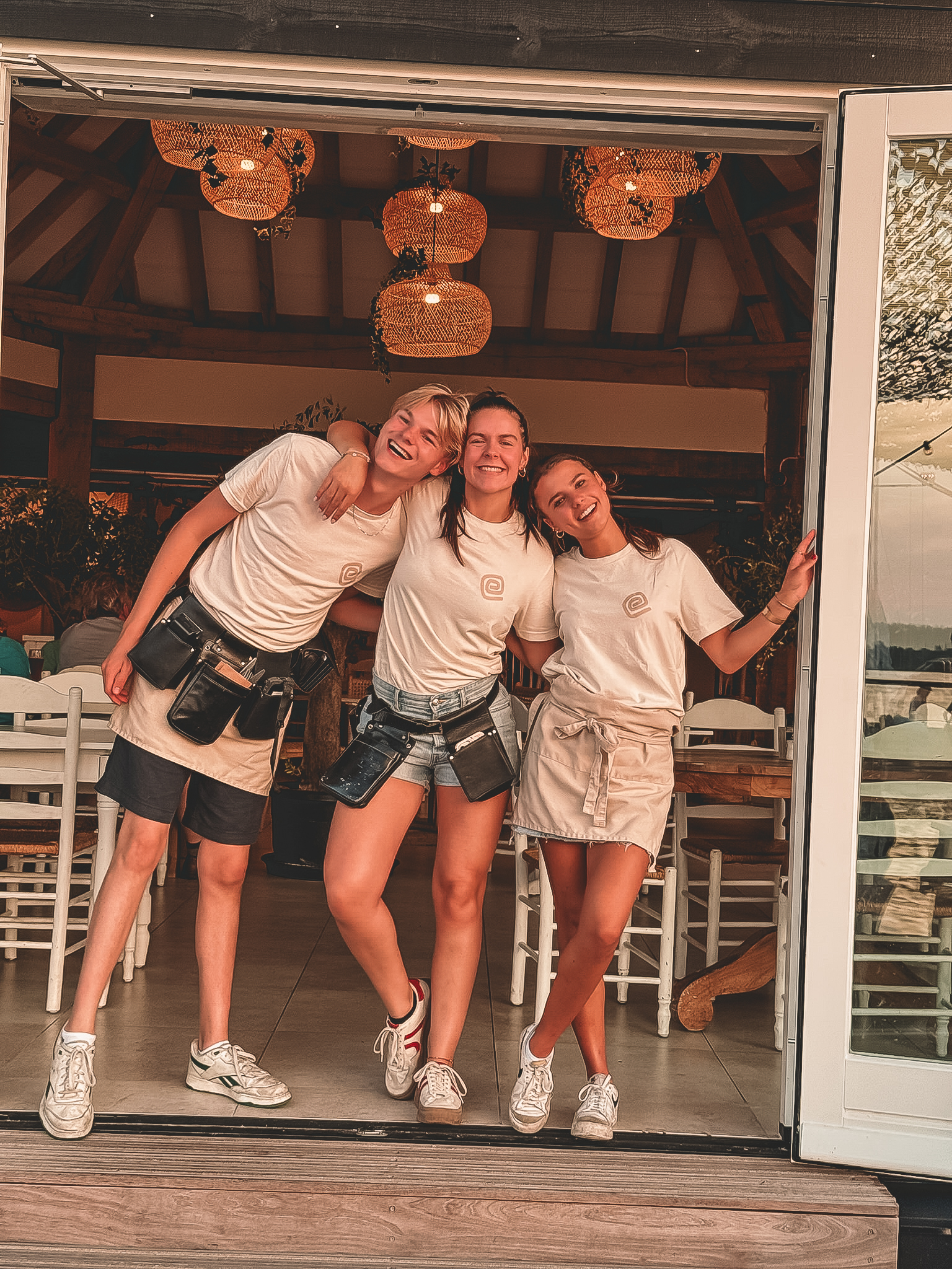 Drie lachende medewerkers in casual uniformen, omarmd in een gezellige restaurantomgeving met houten decor.