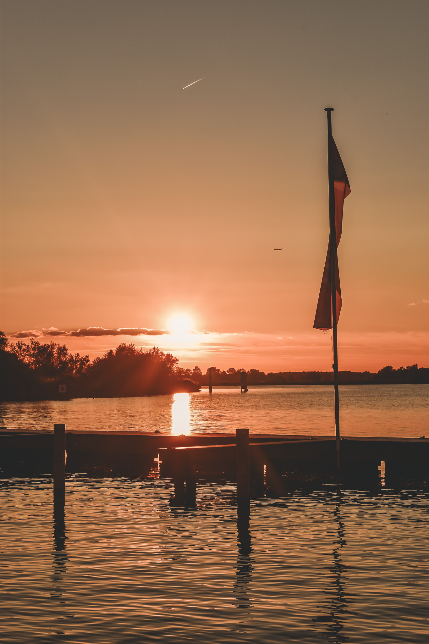 Zonsondergang boven een rustige rivier met silhouet van een vlaggenmast en bomen, warme oranje tinten.
