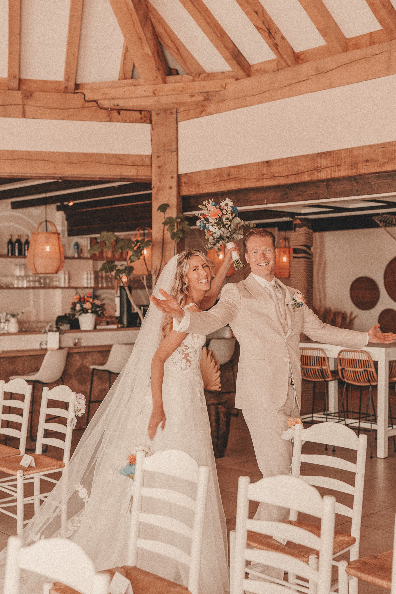Bruidspaar in lichte trouwkleding straalt in een sfeervolle, houten trouwlocatie met bloemen en decor.