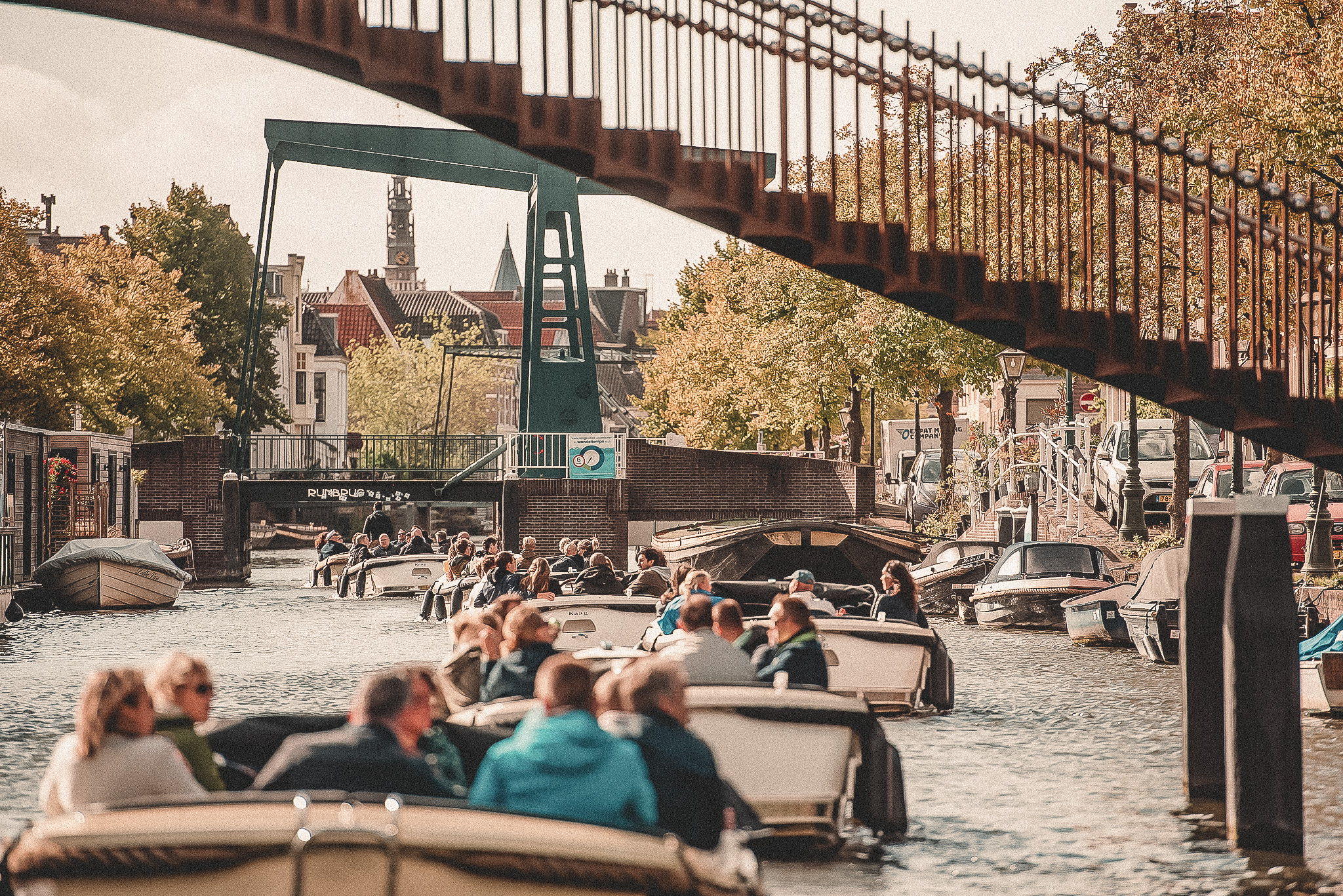 Boten vol mensen varen door een zonovergoten kanaal onder een brug, omringd door herfstkleurige bomen.