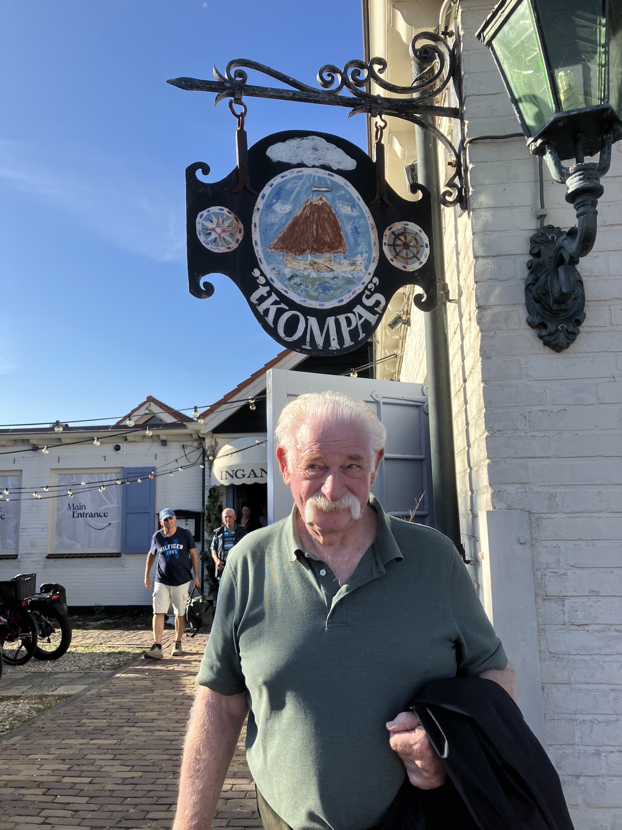 Man voor restaurant 't Kompas, met nostalgisch uithangbord en zonnige blauwe lucht op achtergrond.