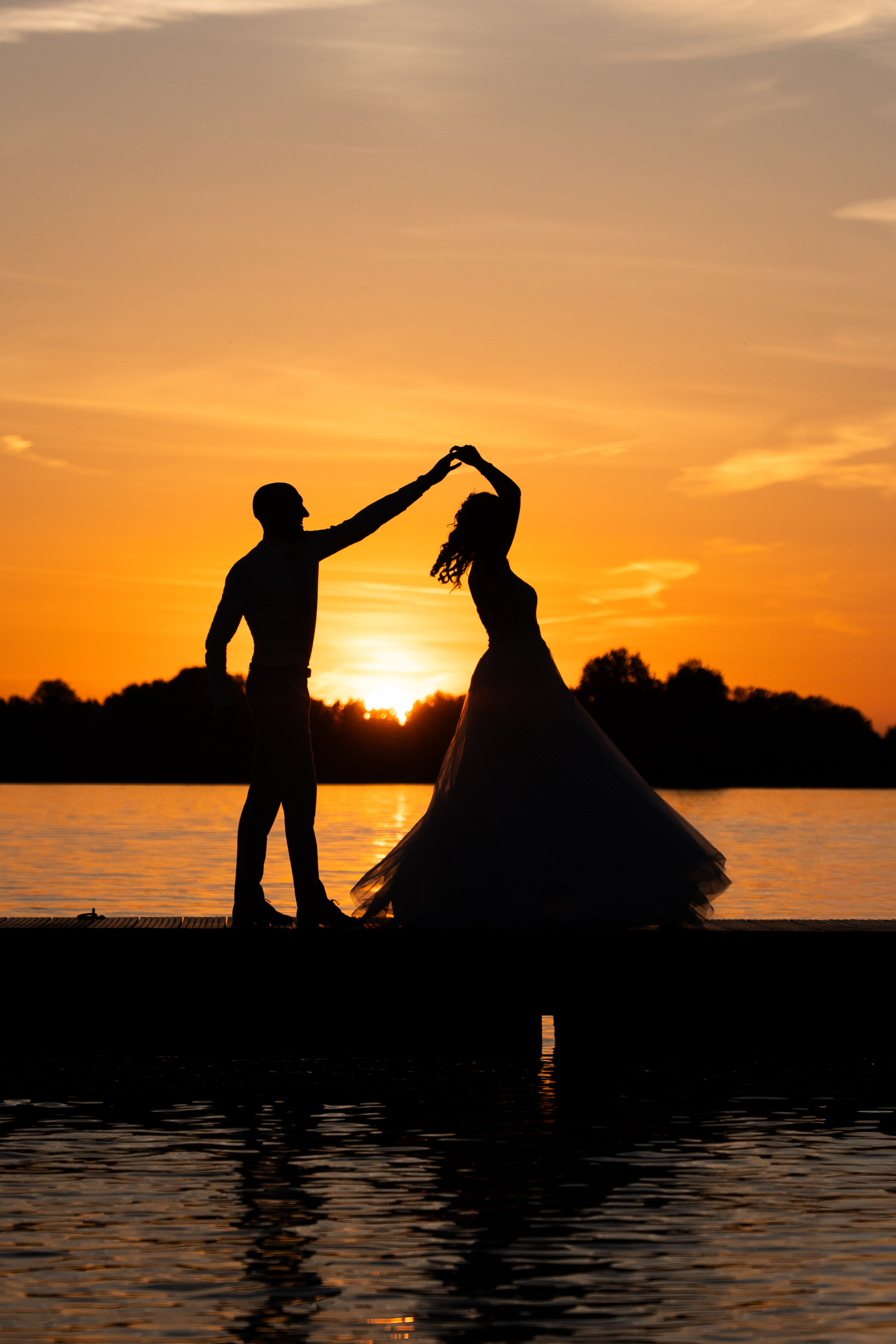 Twee silhouetten dansen op een steiger bij zonsondergang, met een oranje lucht en weerspiegeling in het water.