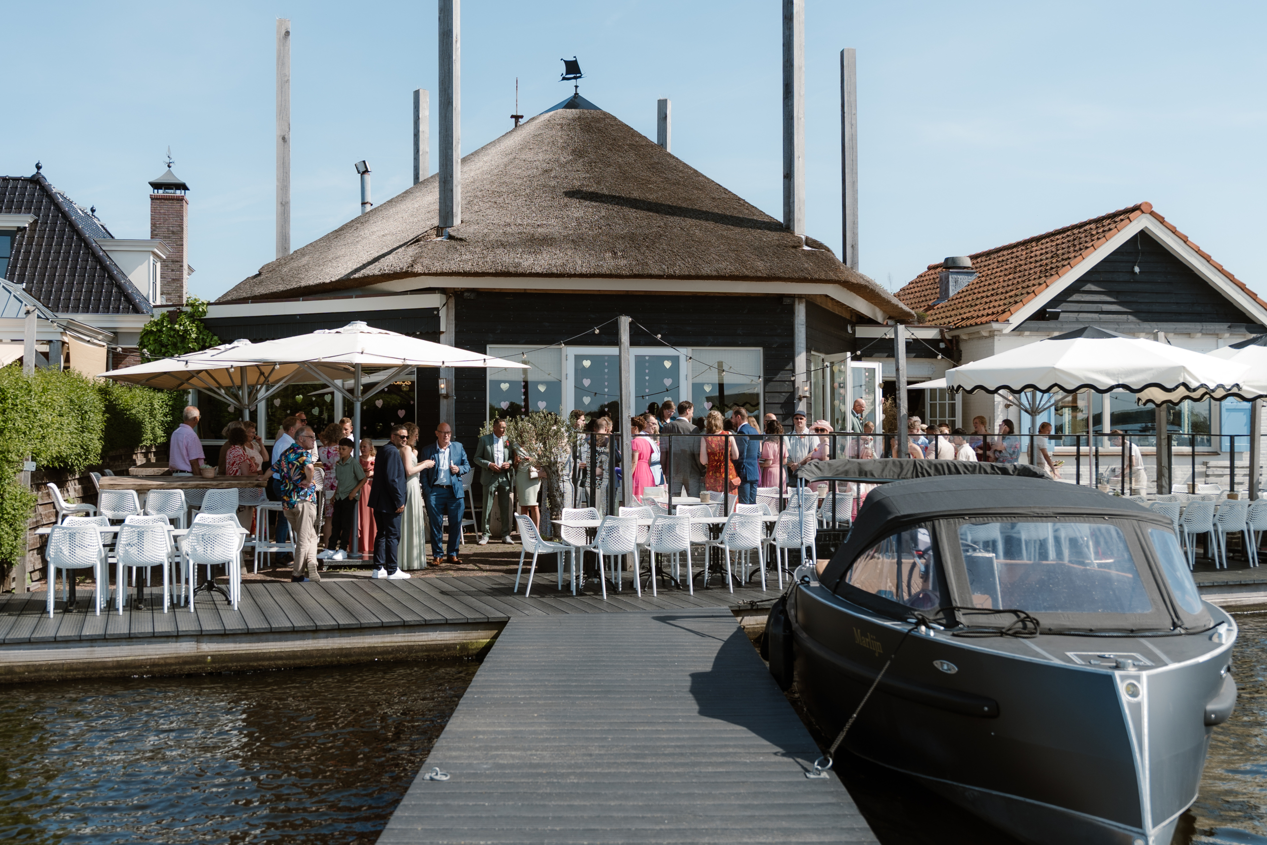 Gezellige buitenbijeenkomst bij rietgedekte locatie aan het water, met gasten onder witte parasols.