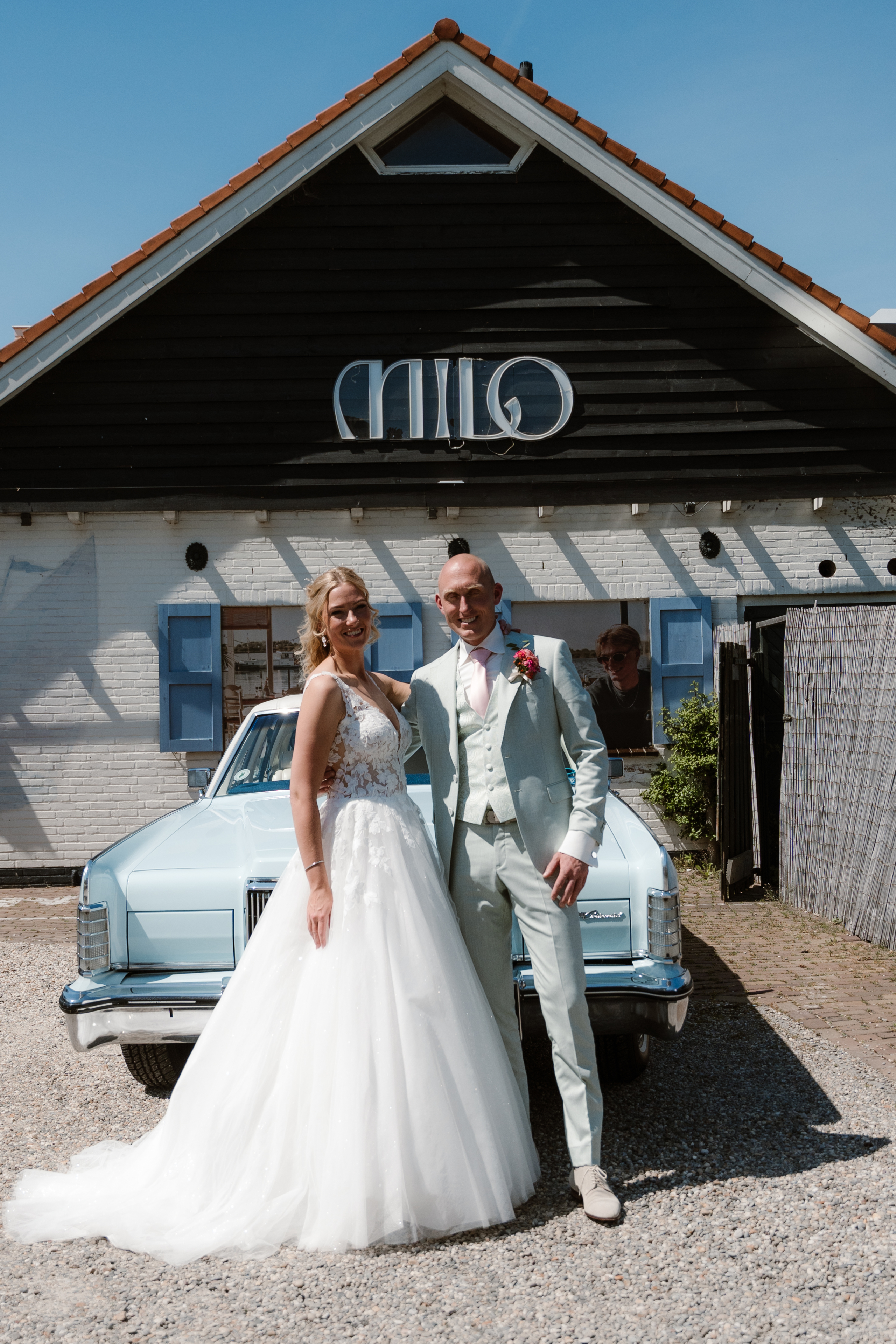 Bruidspaar poseert voor een lichtblauwe vintage auto, voor een gebouw met een zwarte gevel en blauwe luiken.