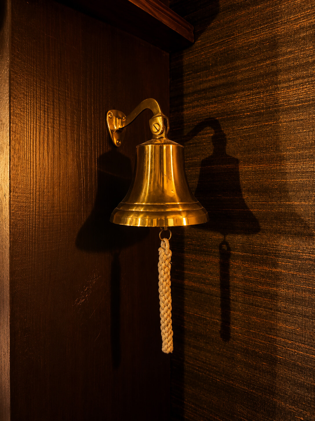 Messing bel met gevlochten touw, bevestigd aan een donkere houten muur, warme sfeerverlichting.
