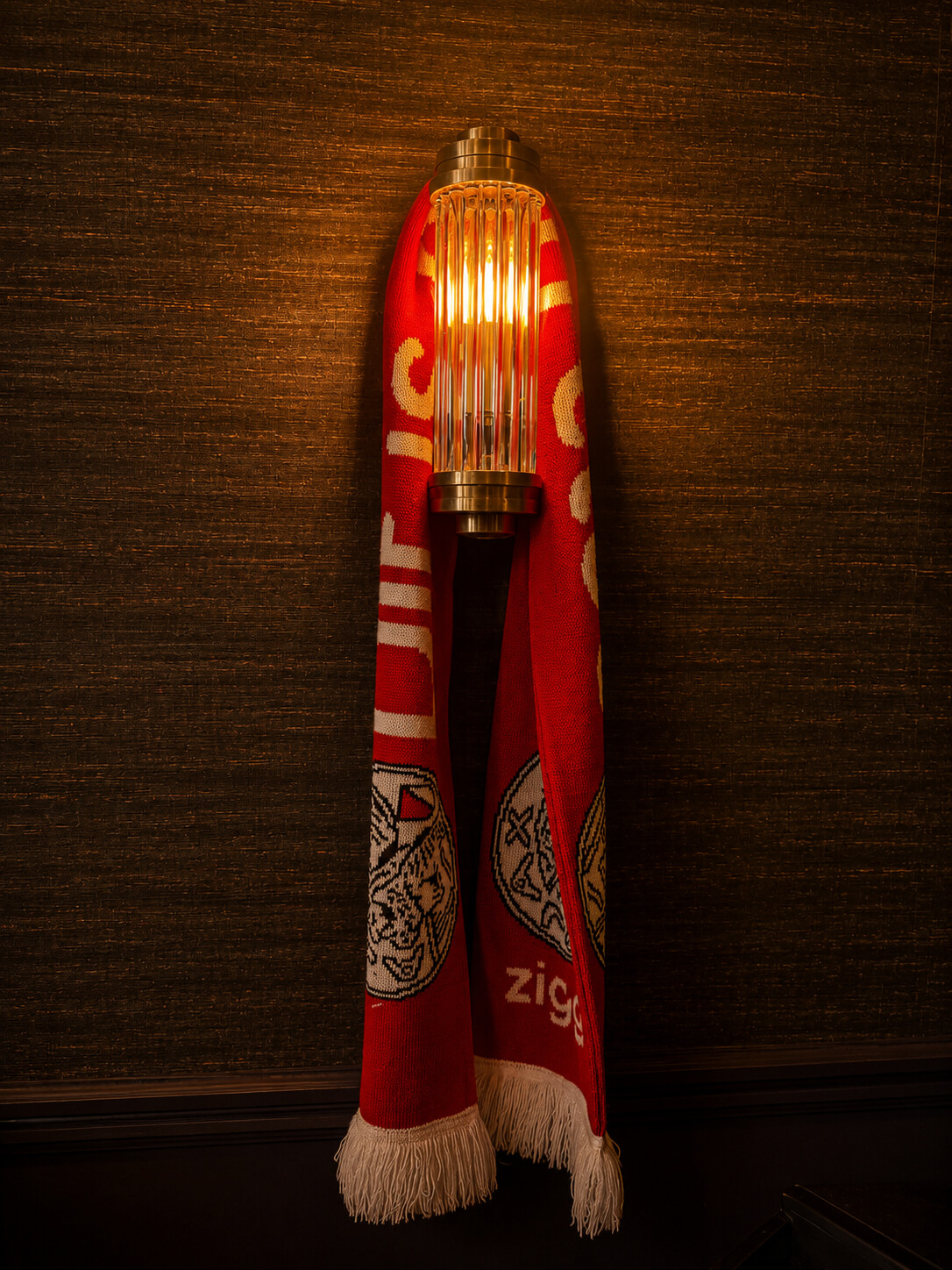 Rode sjaal met wit patroon hangt over een warme, goudkleurige wandlamp op een donkere muur.