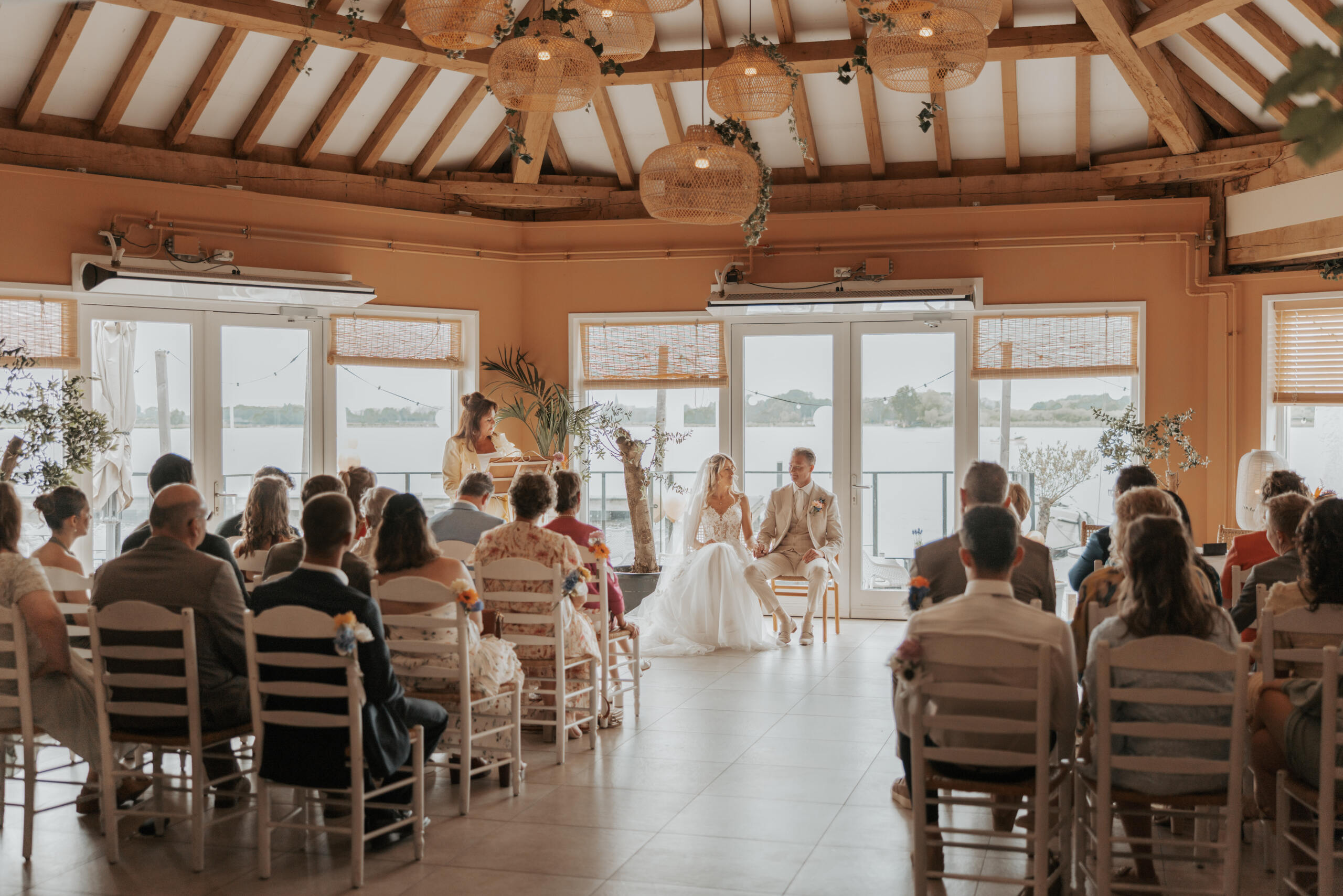 Bruiloftceremonie in lichte, rustieke zaal met houten balken, rieten lampen en uitzicht op het water.