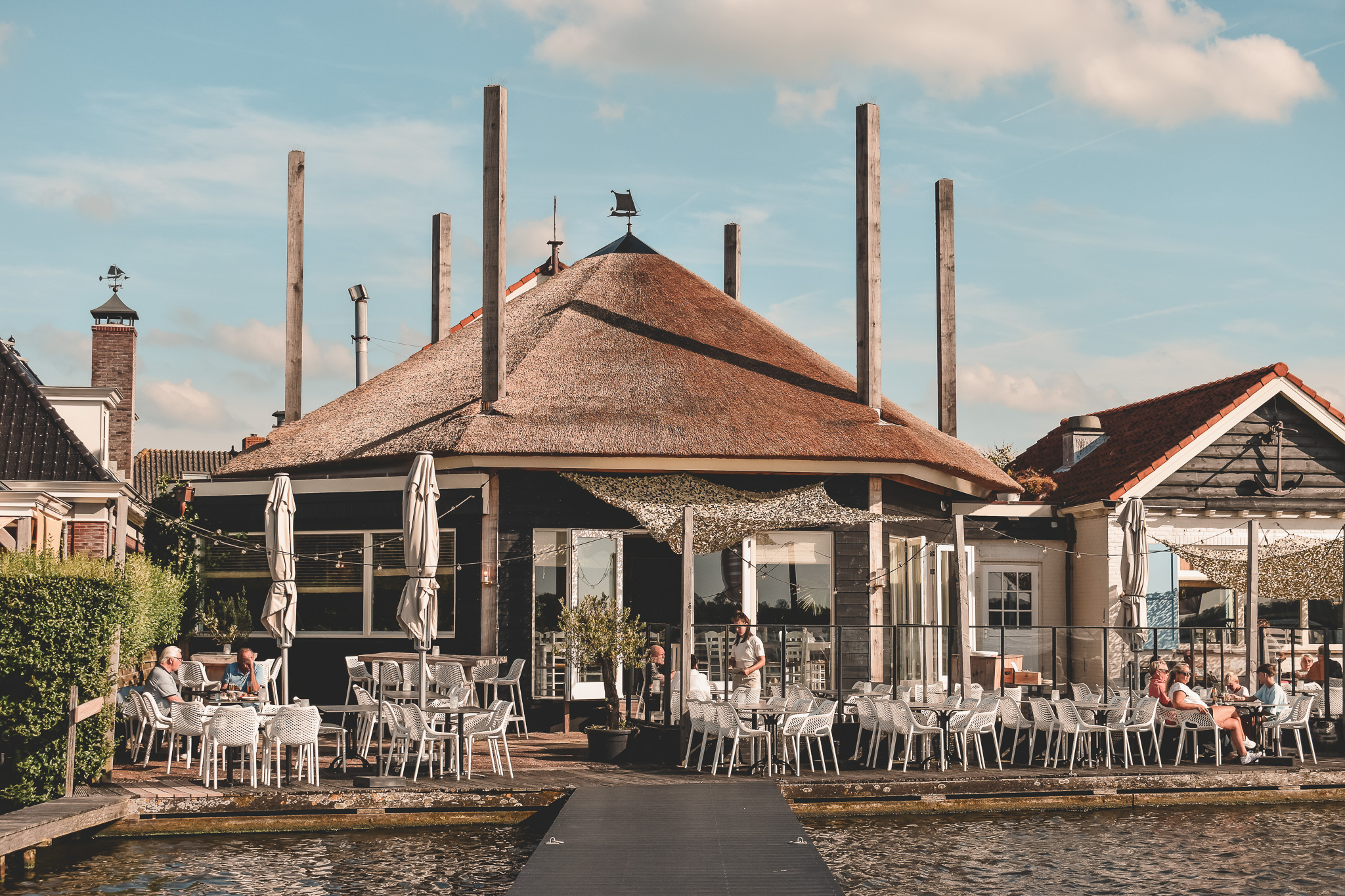 Rietgedekte restaurant aan het water met terras vol witte stoelen, zonnige sfeer en blauwe lucht.