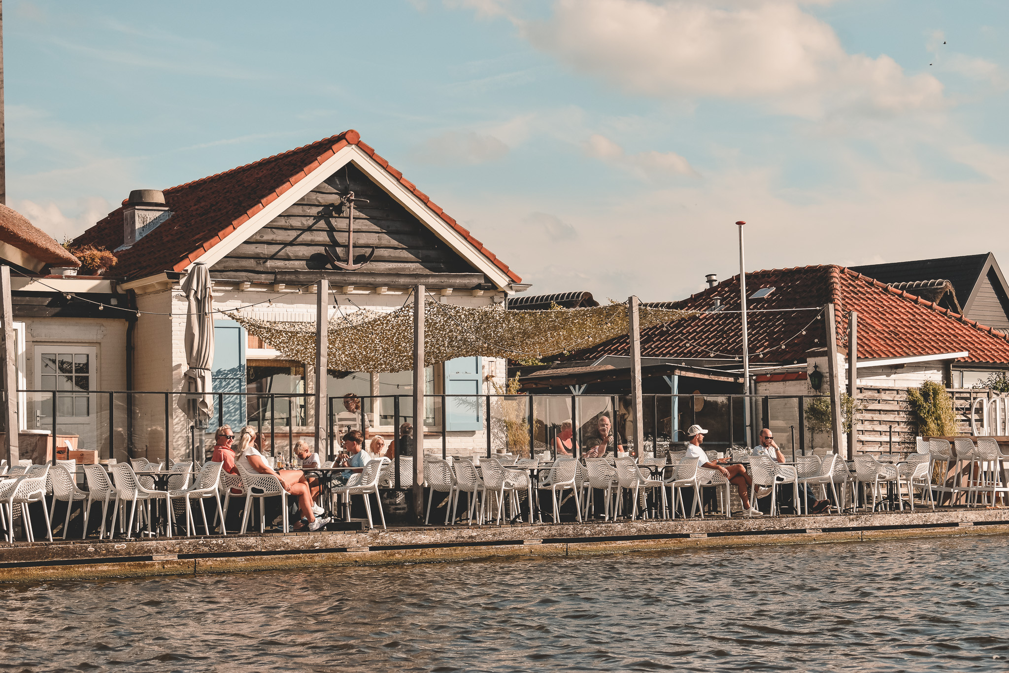 Terras aan het water met witte stoelen, rustieke houten gebouwen en decoratieve overkapping. Zonnige sfeer.