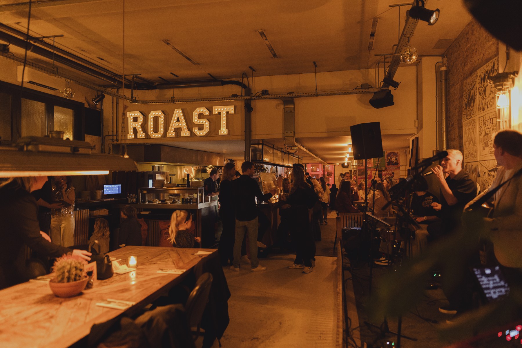 Gezellige bar met warme verlichting, houten tafels en een bord met "ROAST". Mensen genieten van de sfeer.