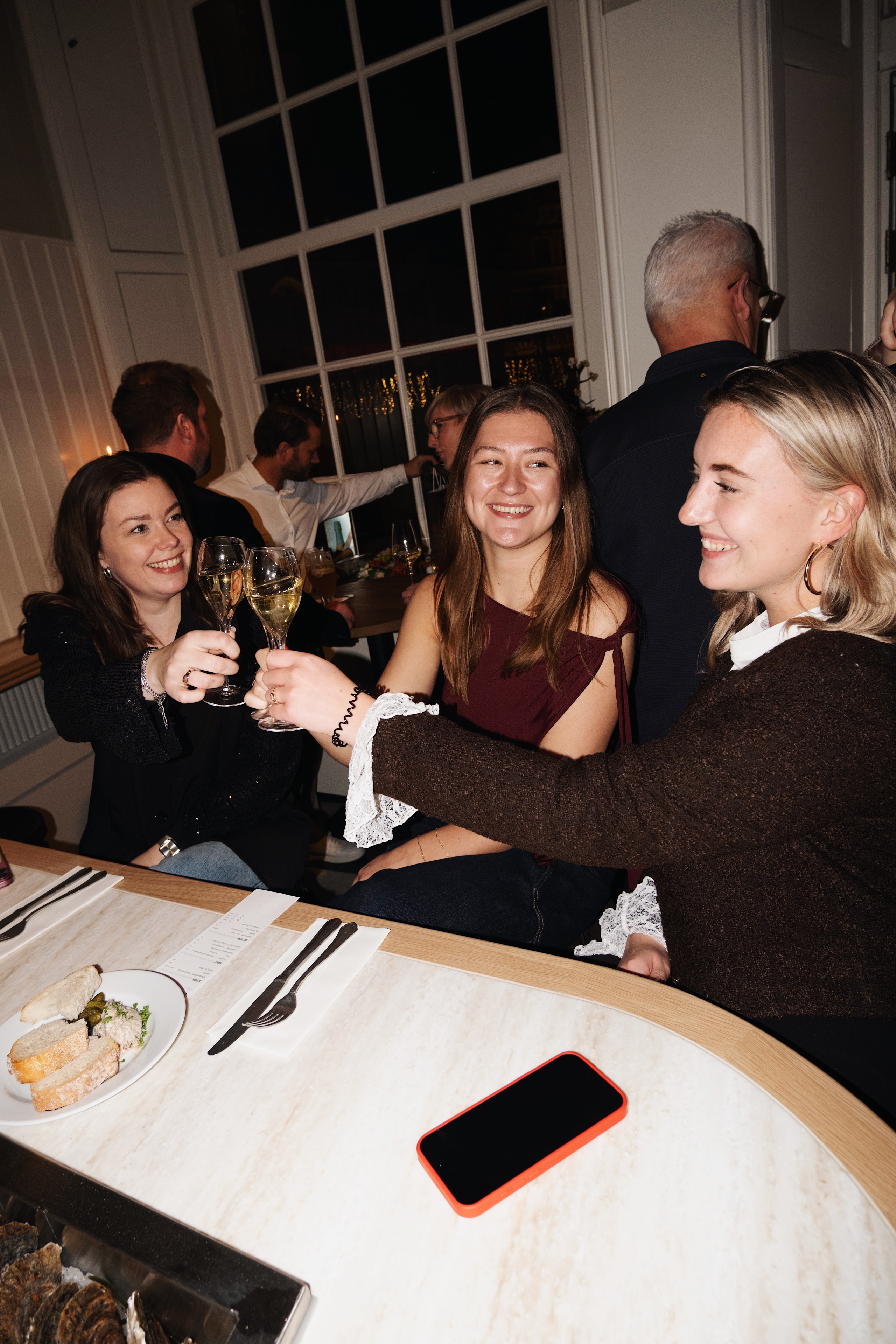 Drie vrouwen proosten met champagneglazen, zittend aan een houten tafel in een gezellige restaurantomgeving.