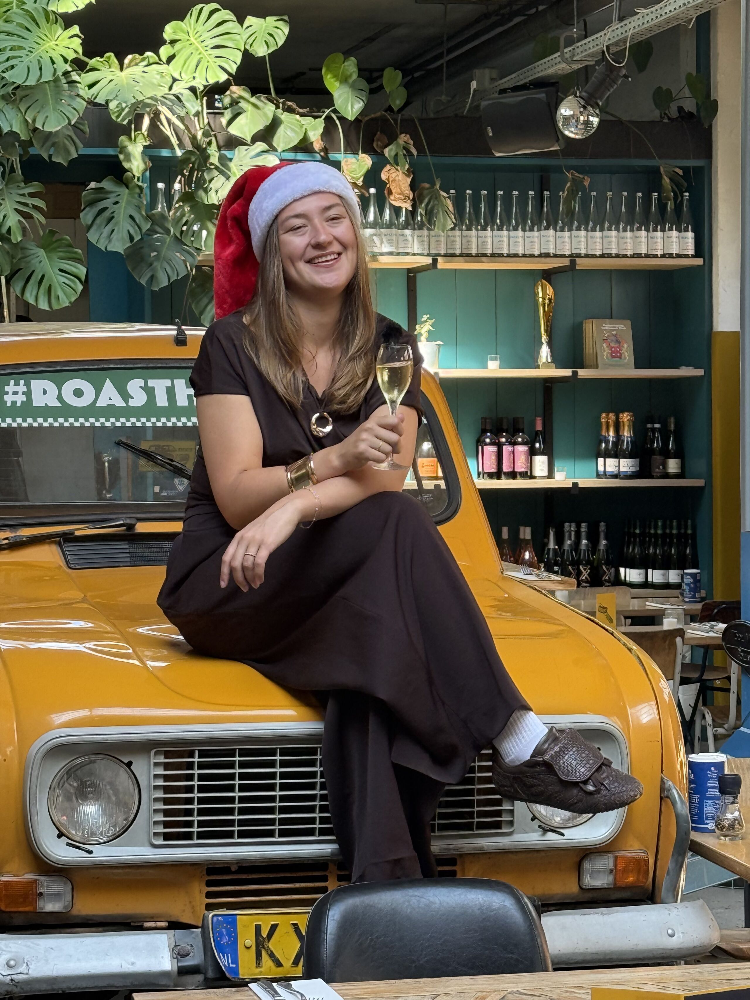 Vrouw in kerstmanmuts zit op gele auto met glas champagne, omringd door groene planten in een gezellige bar.