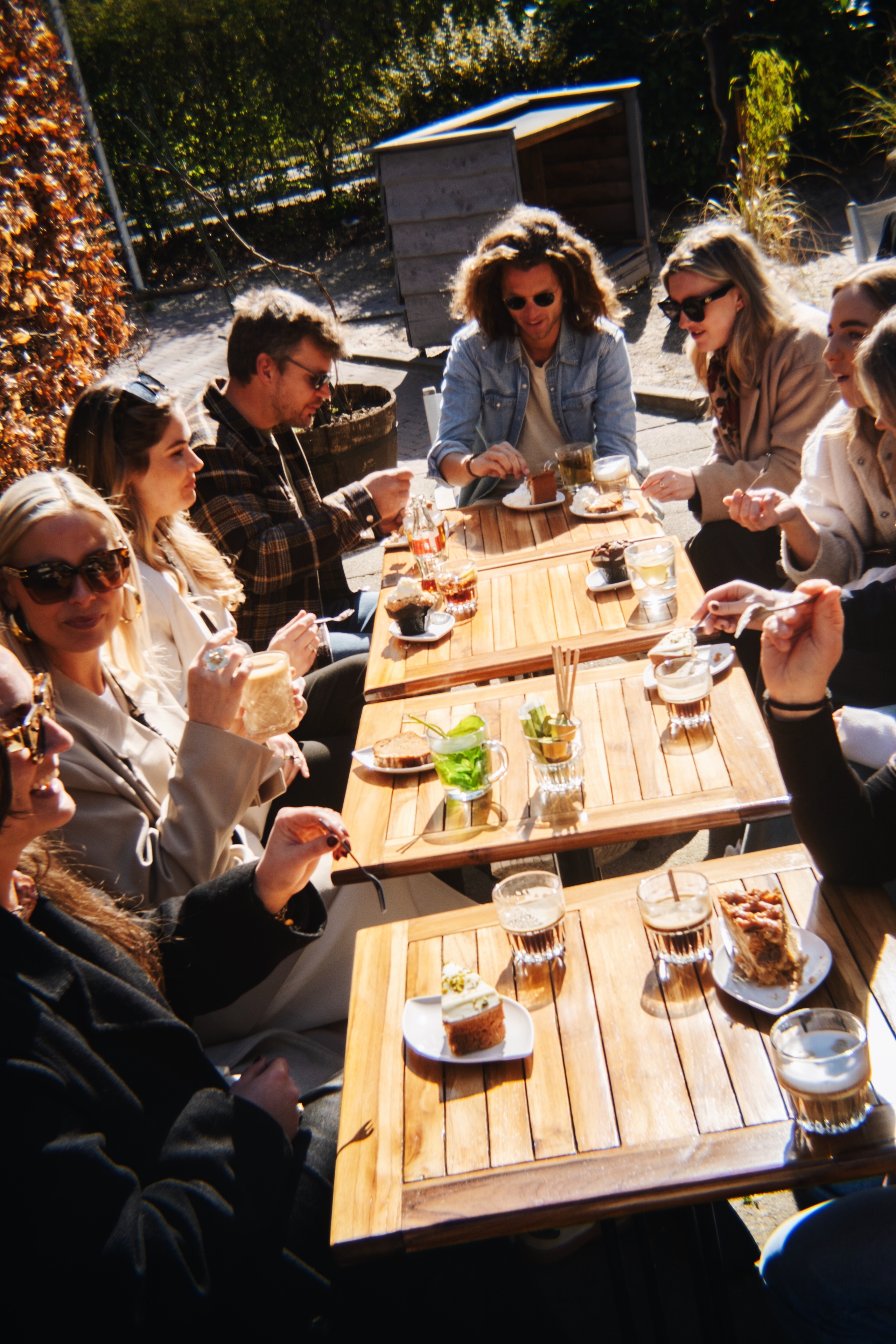 Groep vrienden geniet van koffie en taart aan houten tafels in een zonnige buitenomgeving.
