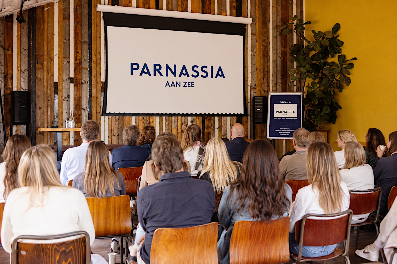 Publiek zit voor een projectiescherm met "Parnassia aan Zee", houten muren en gele wand.