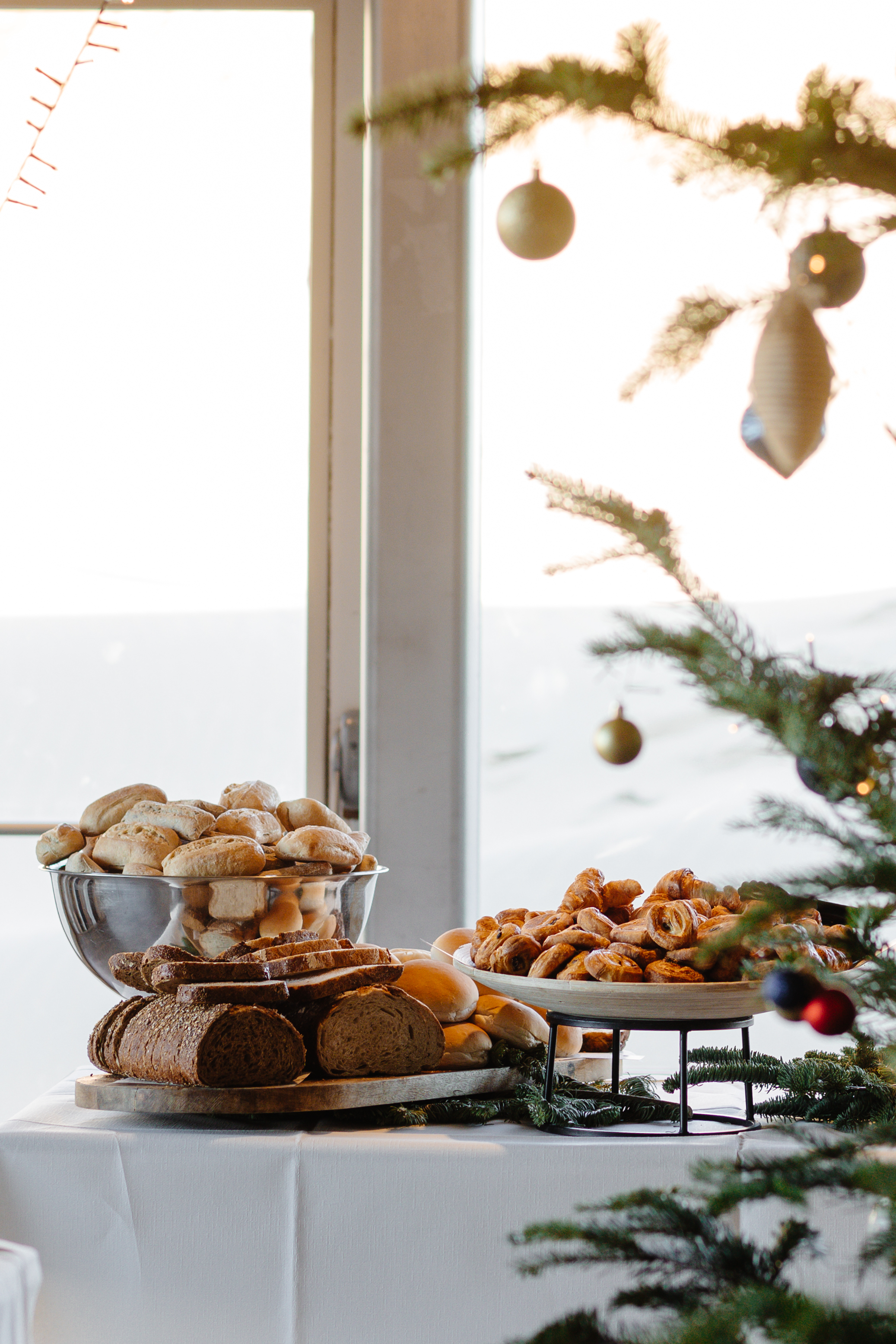 Assortiment van brood en gebak op een wit gedekte tafel, naast een kerstboom met hangende ornamenten.