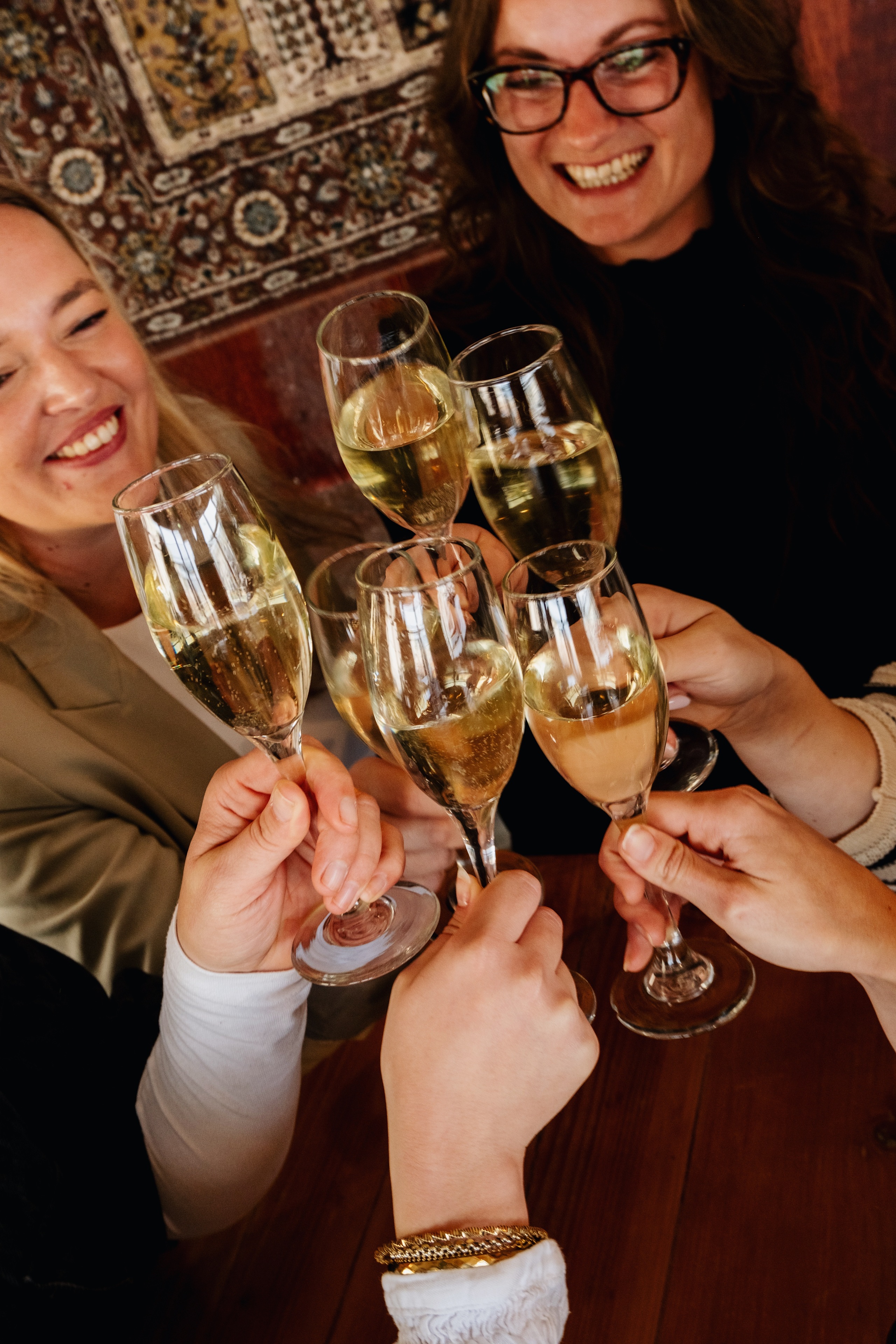 Vrolijke groep mensen proost met glazen champagne, warme sfeer met houten tafel en tapijt op de achtergrond.
