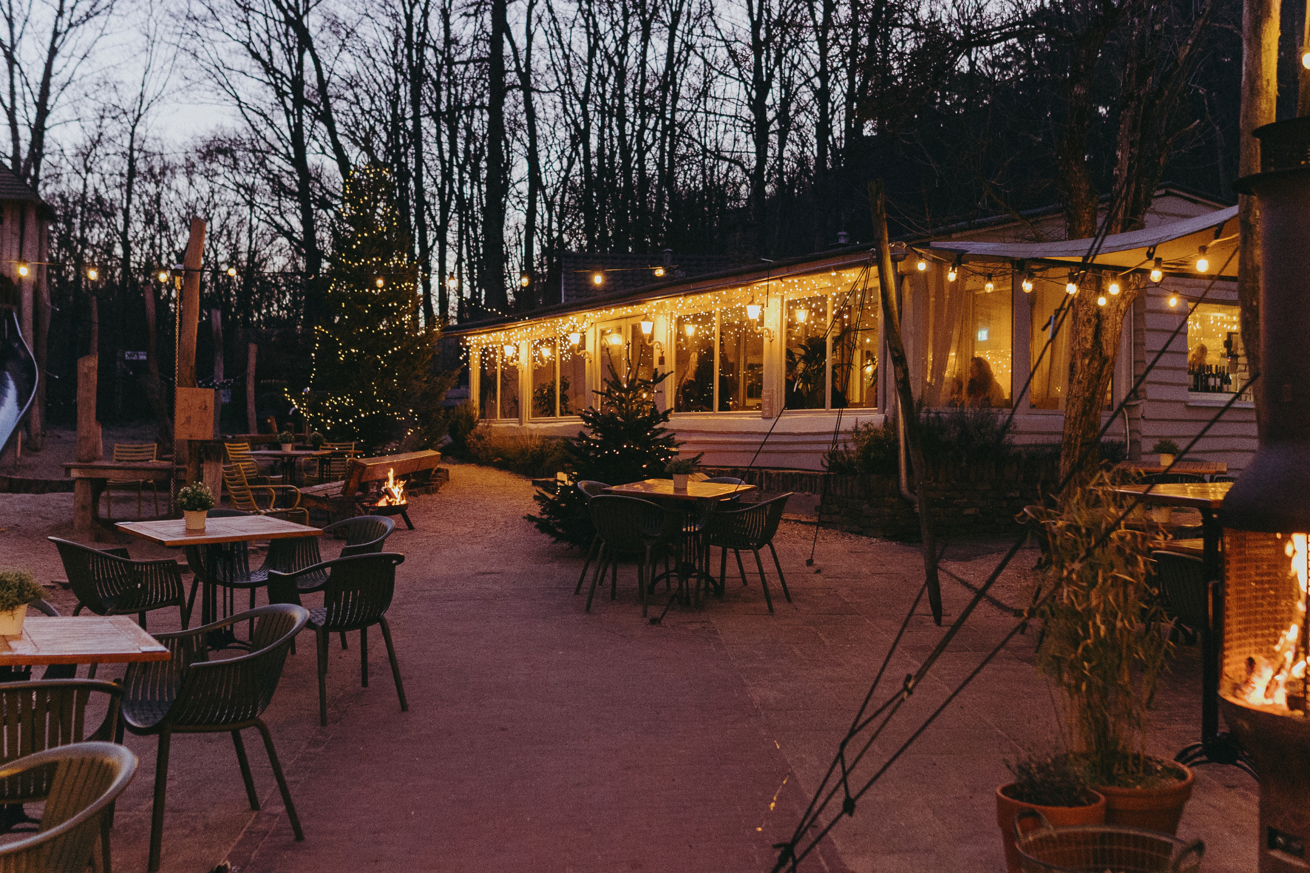Buitenrestaurant met sfeerverlichting, kerstbomen en brandende vuurkorf in een bosrijke omgeving bij schemering.