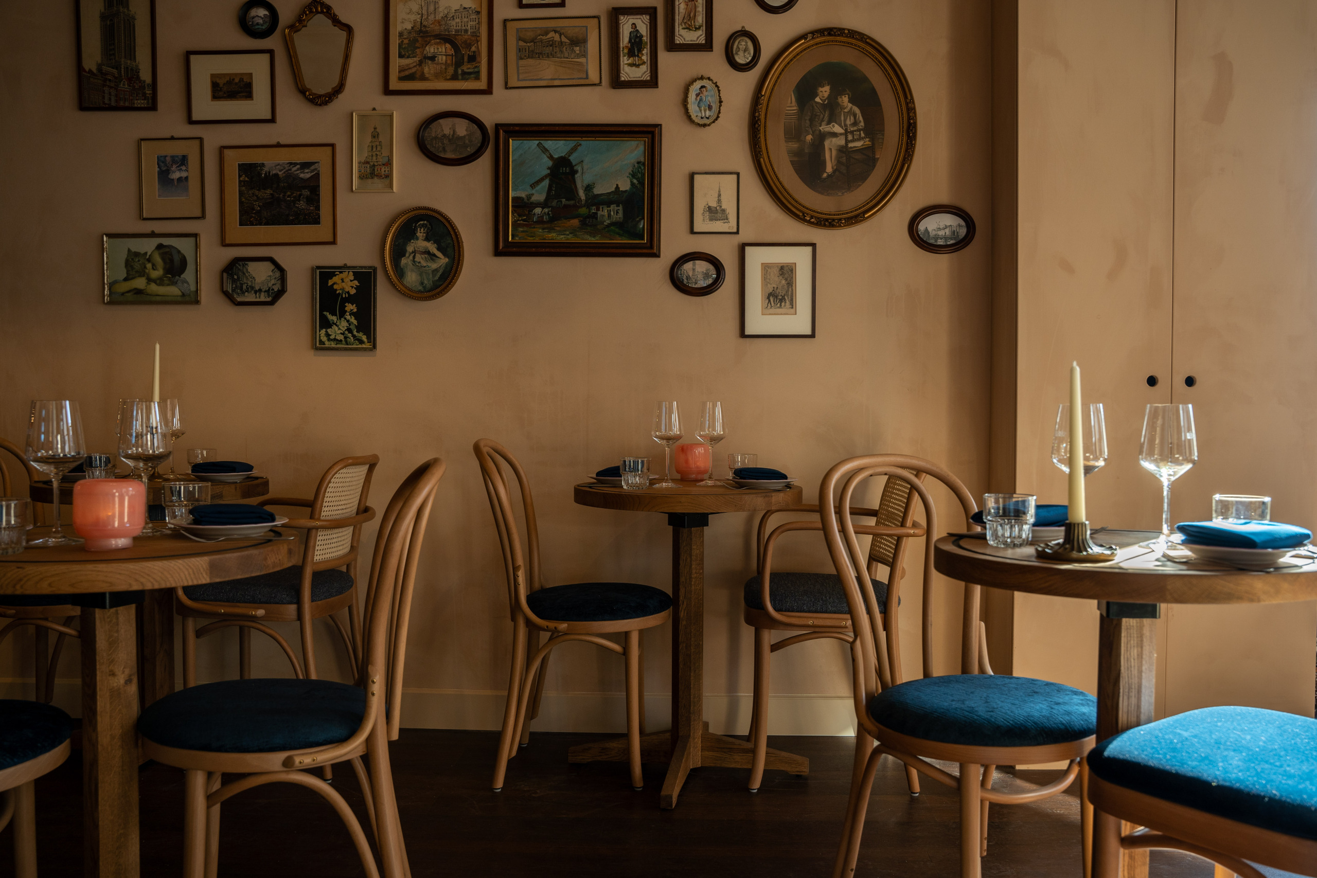 Intieme eetkamer met houten tafels, blauwe stoelen, kaarsen en een muur vol schilderijen in warme tinten.