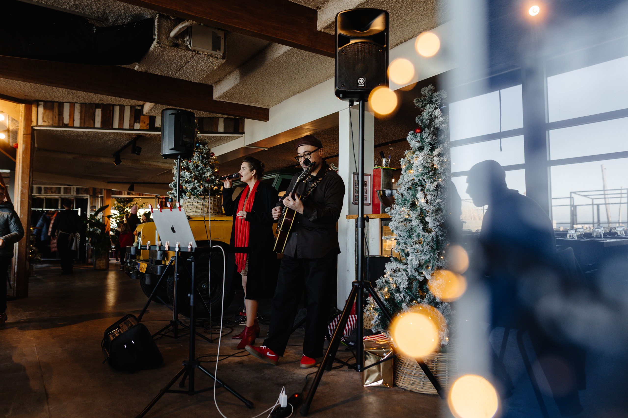 Muzikanten spelen gitaar en zingen naast versierde kerstbomen in een sfeervol restaurant.