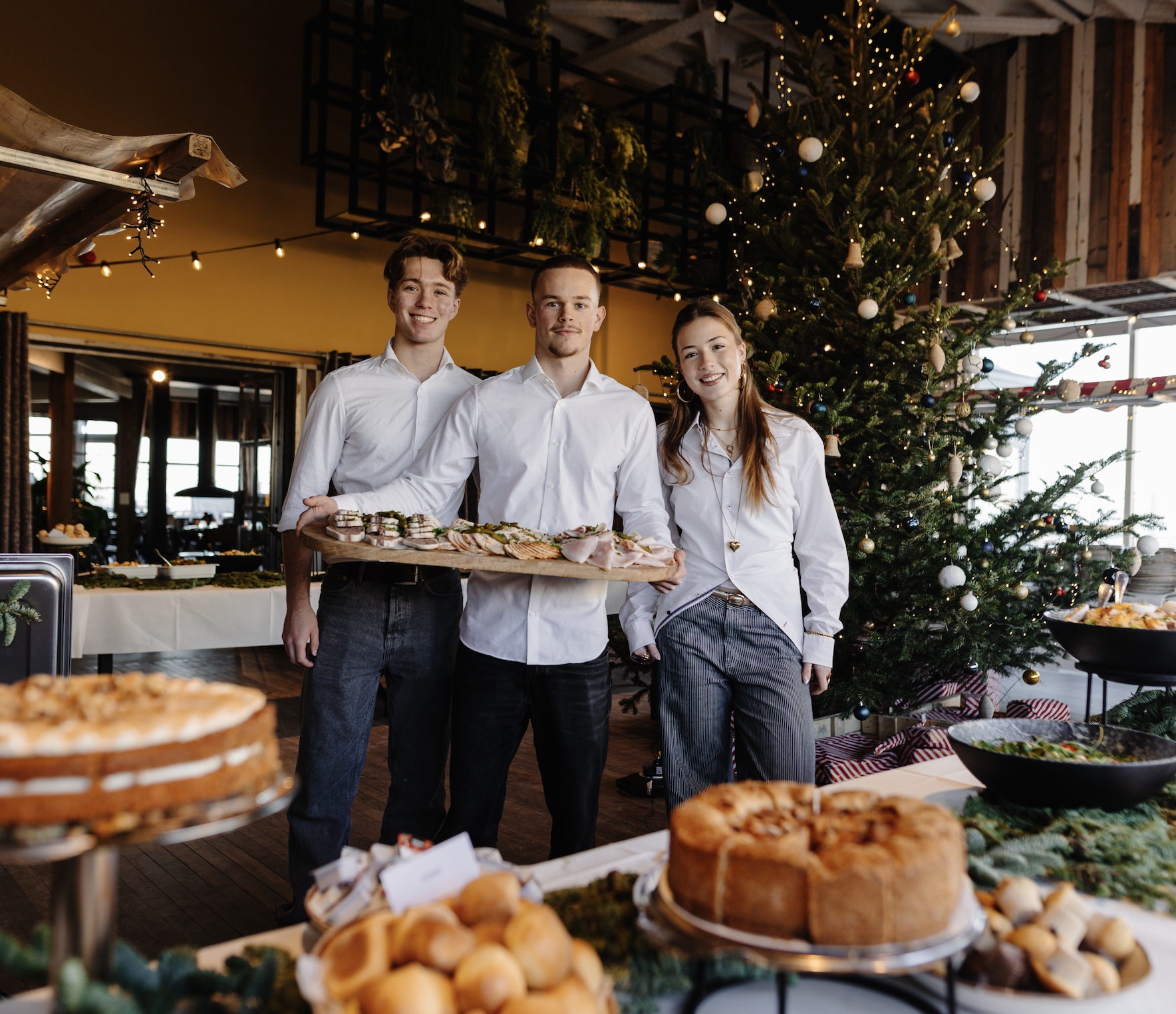 Drie medewerkers in witte overhemden presenteren hapjes voor een versierde kerstboom in een feestelijke ruimte.
