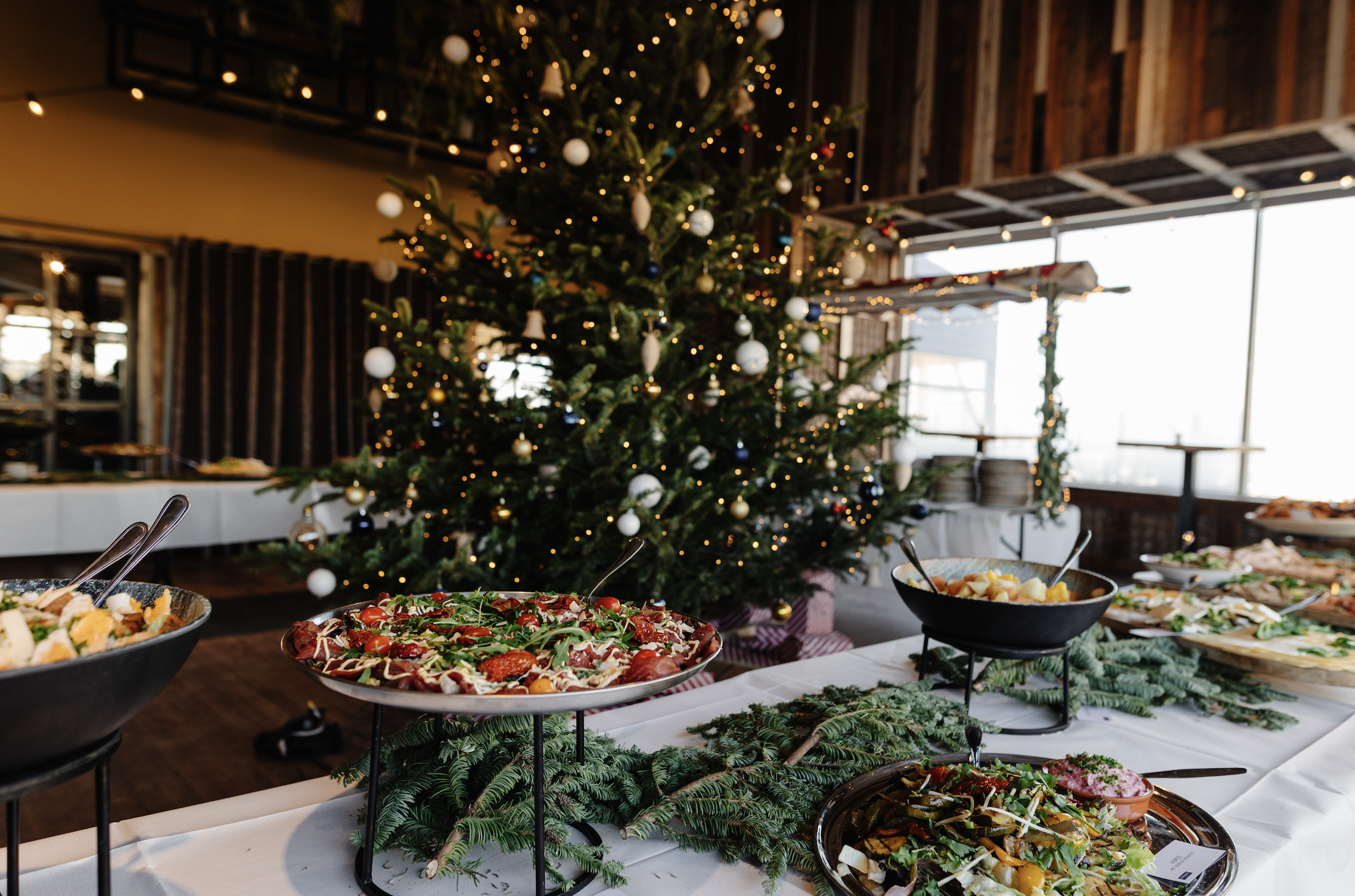 Feestelijke buffettafel met diverse gerechten, kerstboom met lichtjes op de achtergrond, warme sfeer.