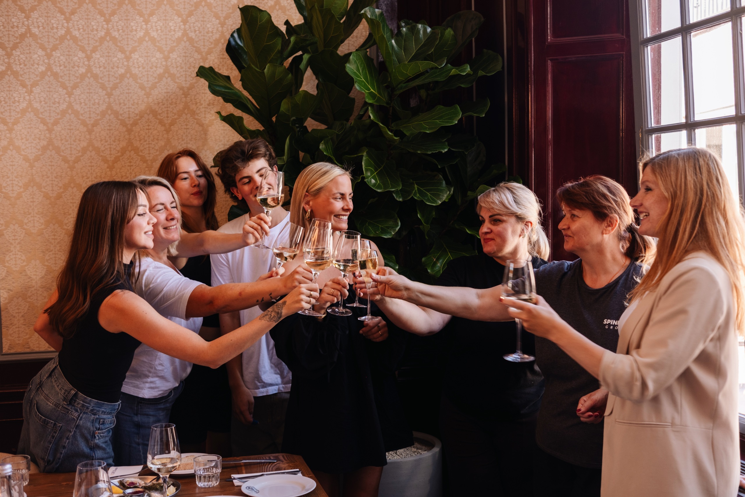 Groep mensen proost met glazen witte wijn in een gezellige, sfeervolle ruimte met planten en warme verlichting.