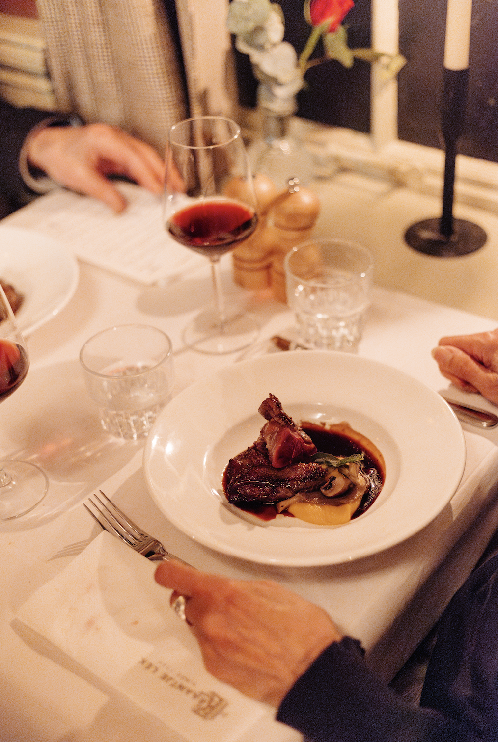 Elegant diner met rood vleesgerecht, rode wijn, kaarslicht en bloem op witte tafel in intieme sfeer.