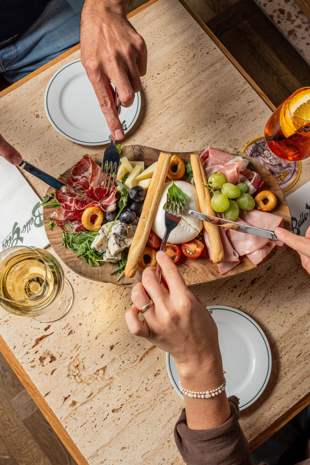 Charcuterie board with meats, cheeses, grapes, and breadsticks on a wooden table, hands reaching in.