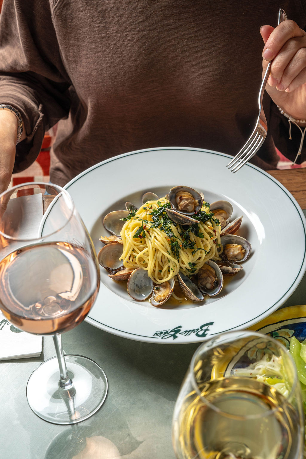 Spaghetti with clams garnished with herbs, paired with a glass of rosé, set on a reflective table.