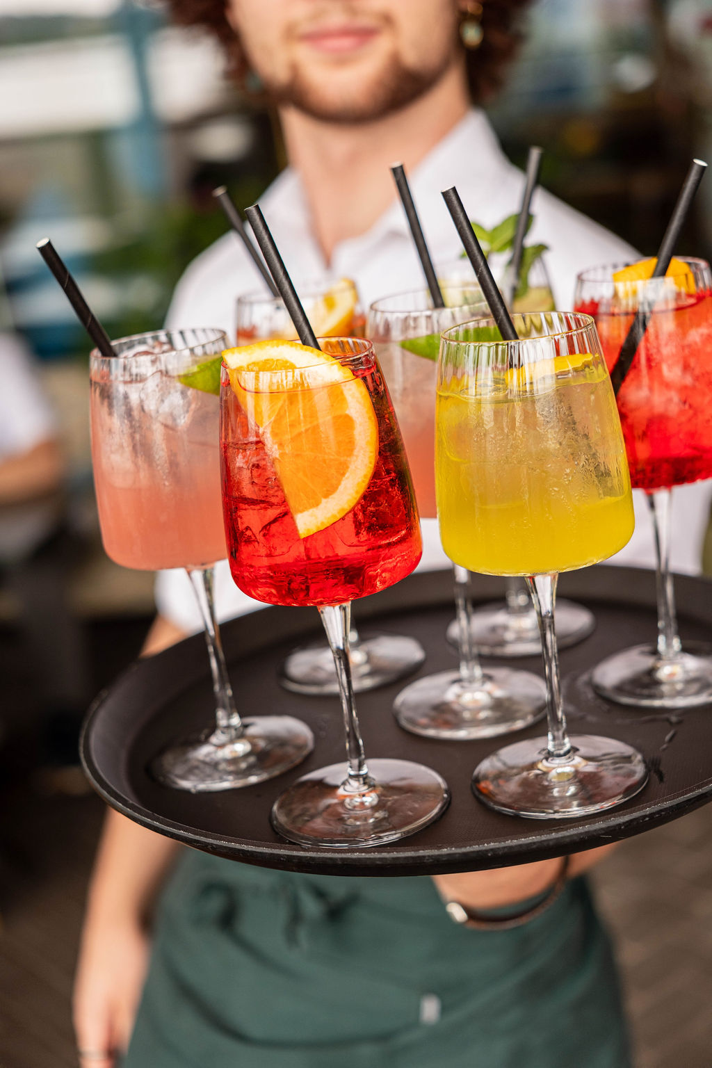 A waiter carries a tray of colorful cocktails with citrus garnishes and black straws, creating a vibrant, refreshing display.
