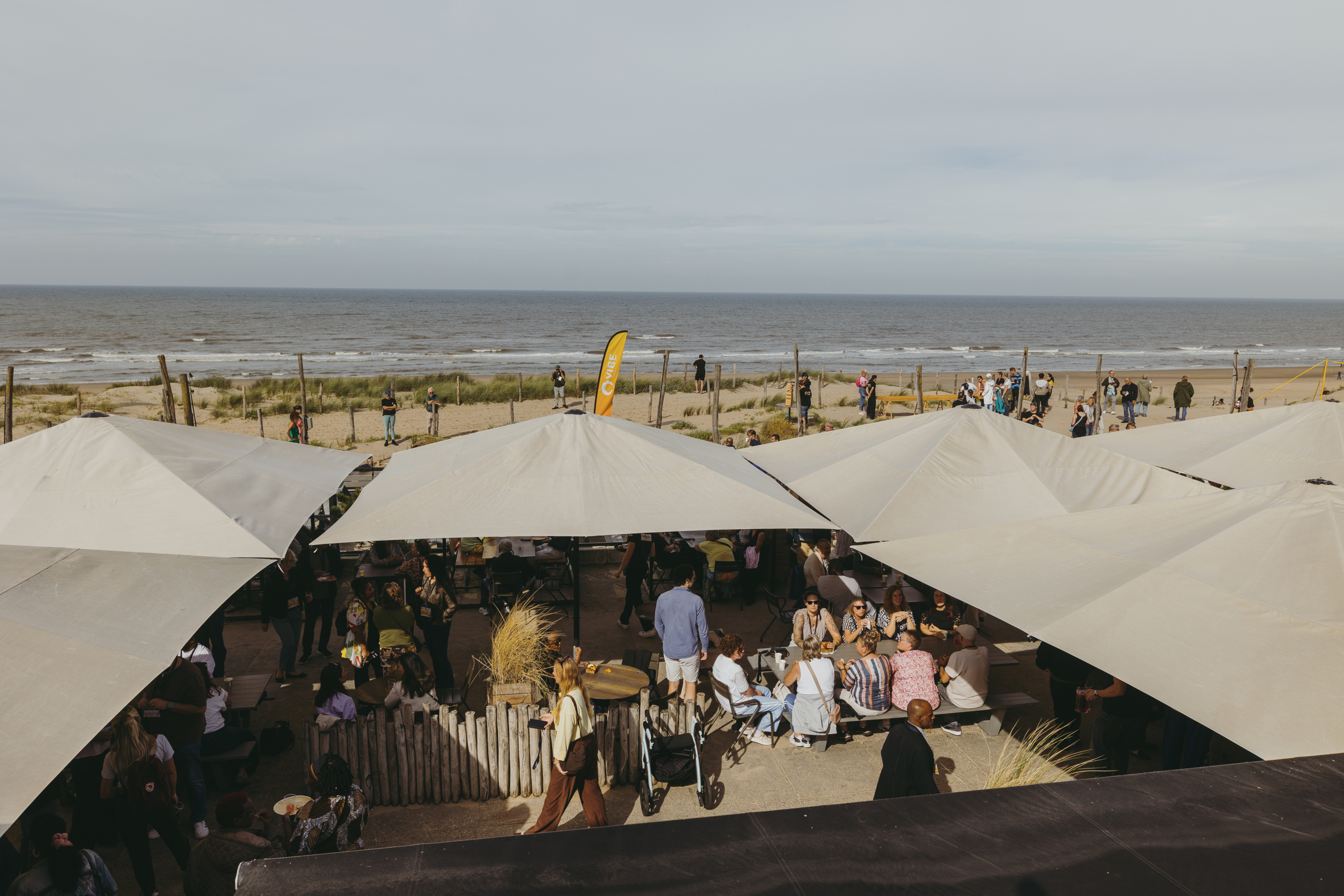 Terras met witte parasols aan het strand, mensen genieten van het uitzicht op zee, ontspannen sfeer.
