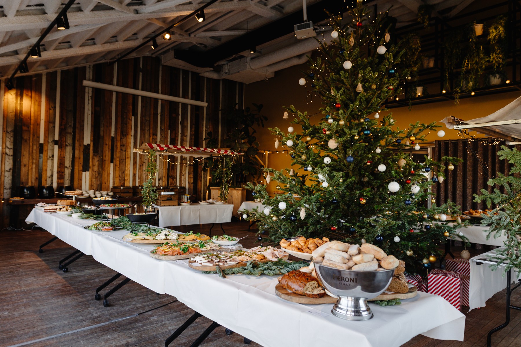 Feestelijke zaal met kerstboom, rijkelijk buffet met diverse gerechten op witte tafelkleden, warme sfeer.
