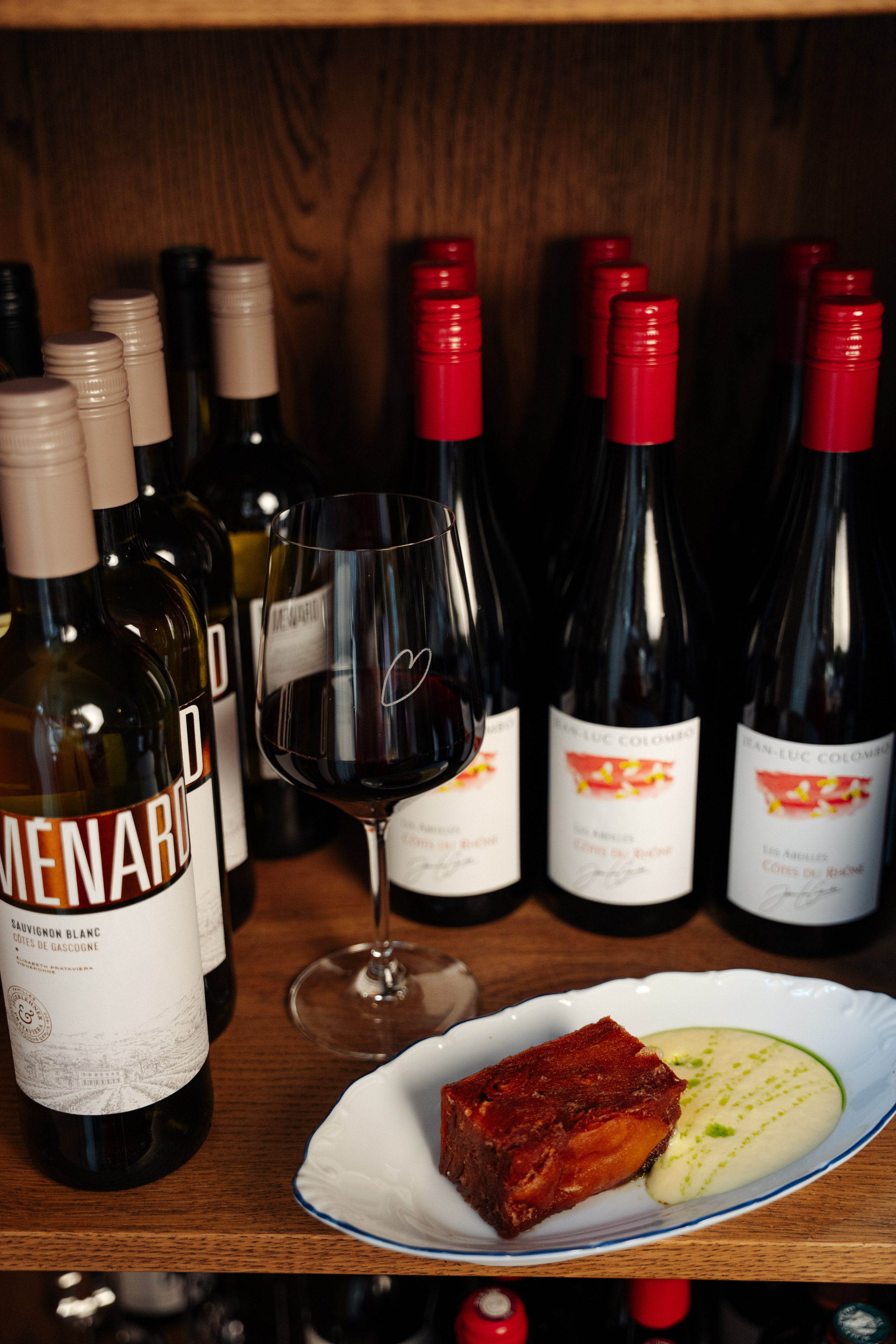 Red wine glass beside a plate of caramelized apple cake with creamy sauce, surrounded by wine bottles.