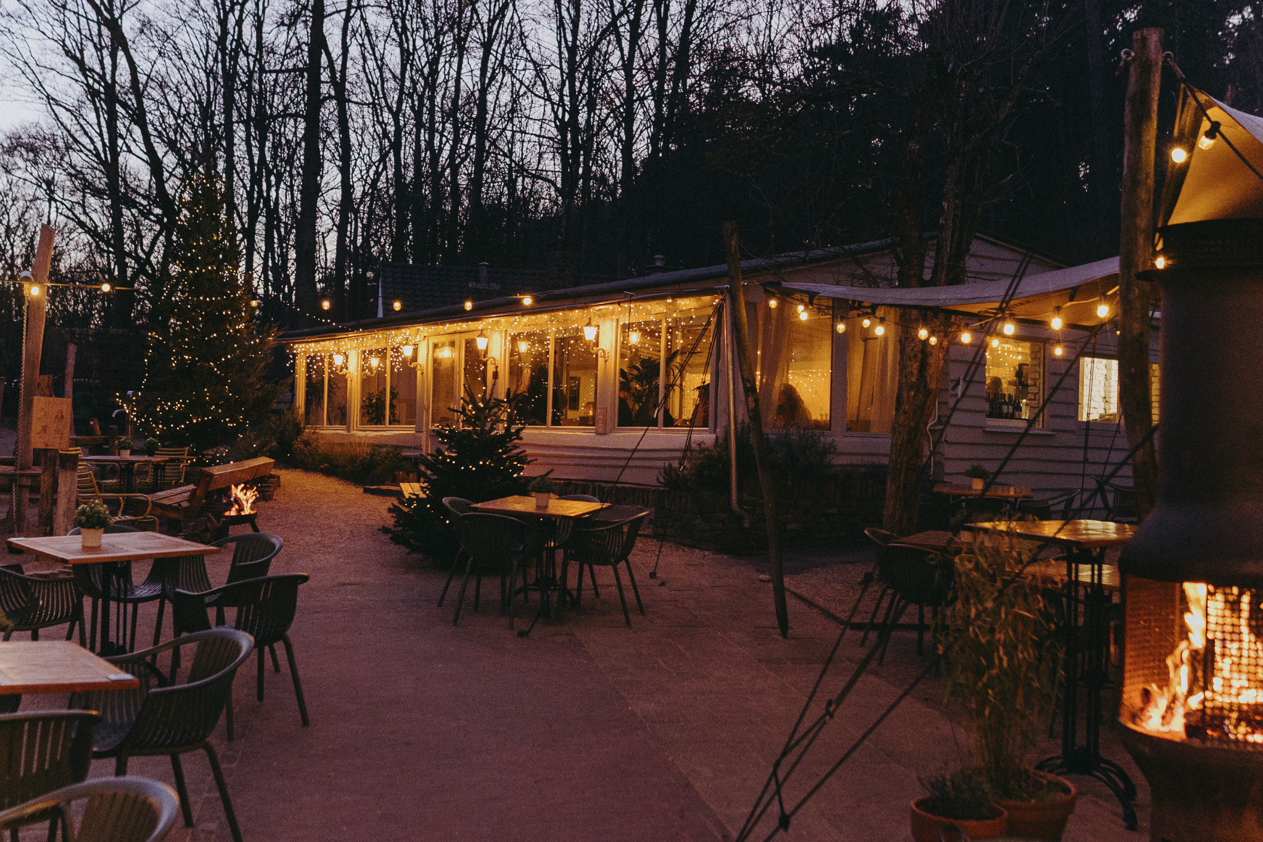 Buitenrestaurant met sfeerverlichting, houten tafels en stoelen. Warme, uitnodigende ambiance bij schemering.