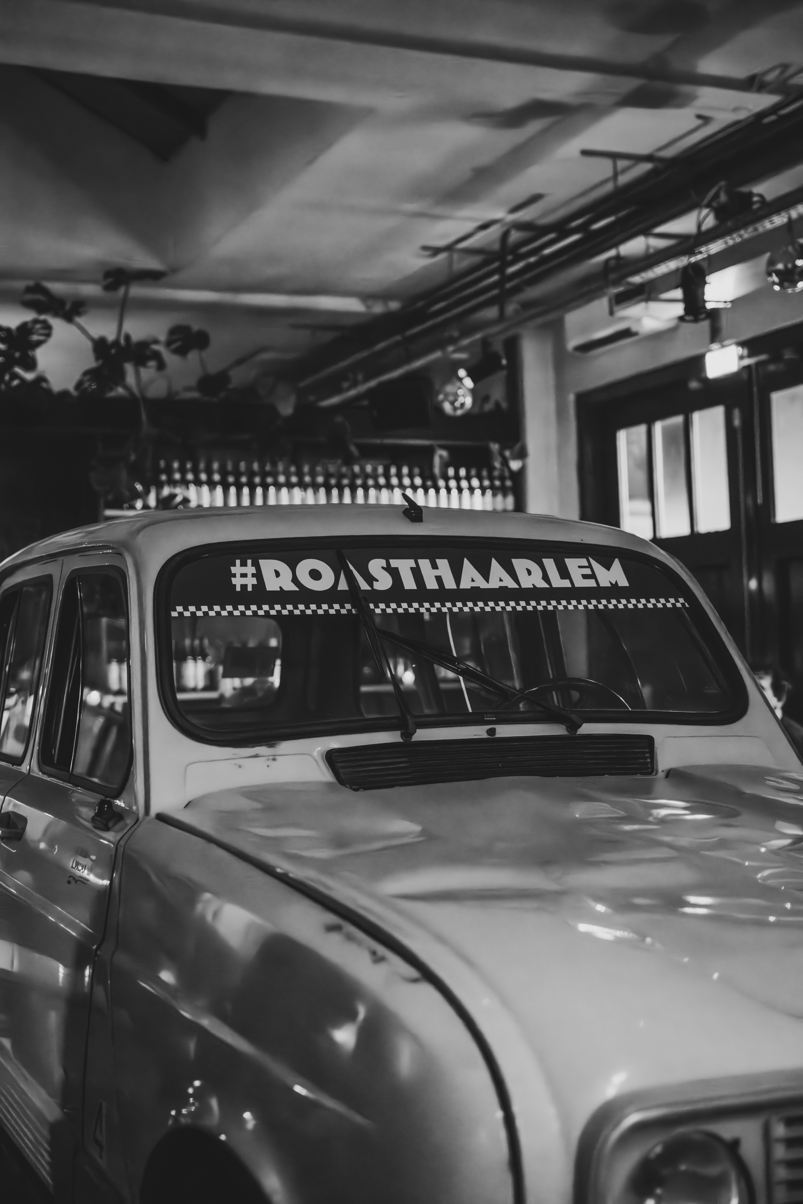 Vintage auto met #ROASTHAARLEM sticker in sfeervol interieur, flessen op achtergrond, zwart-wit.